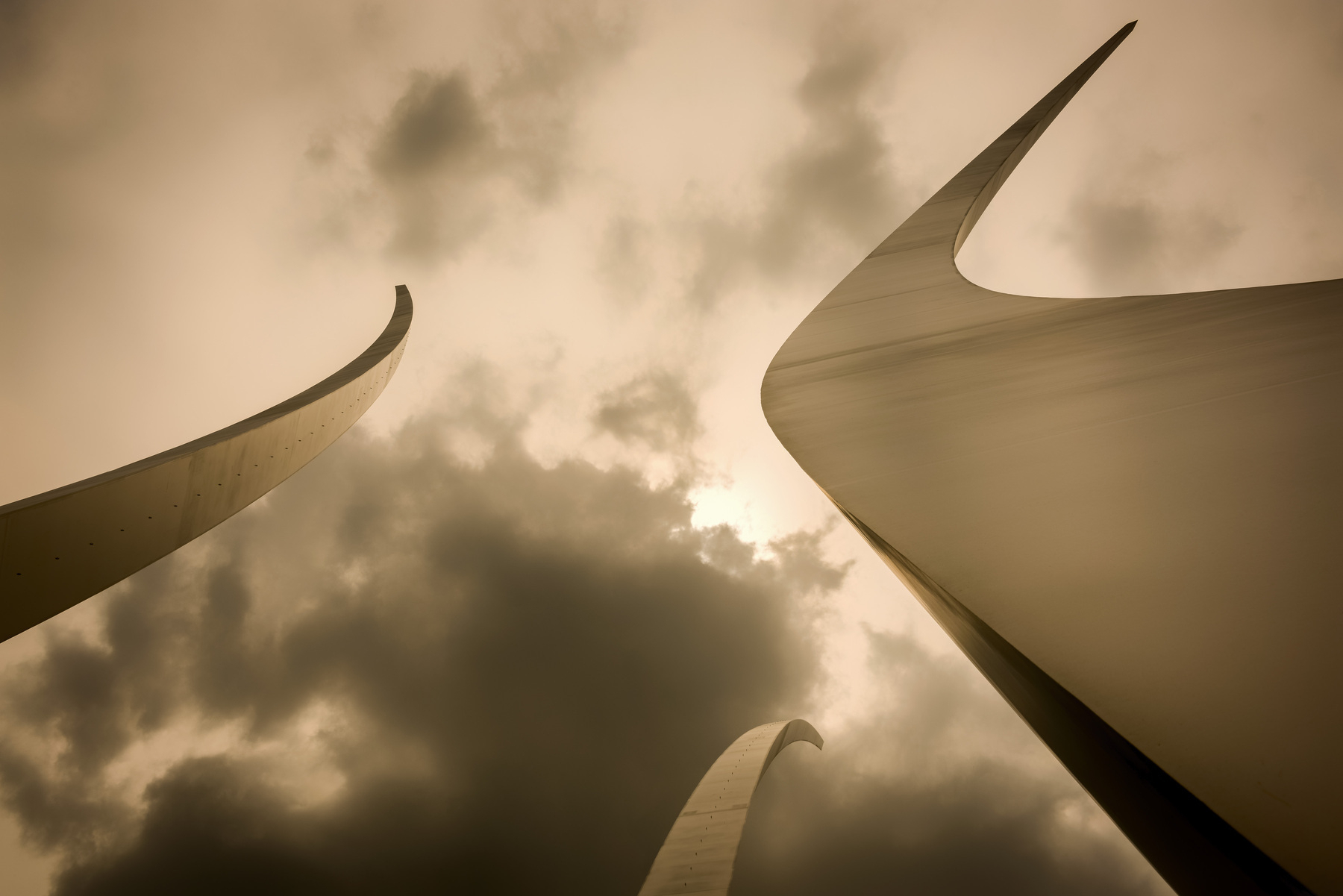 Auto-generated description: Curved, metallic sculptures reach towards a cloudy sky with dramatic lighting.