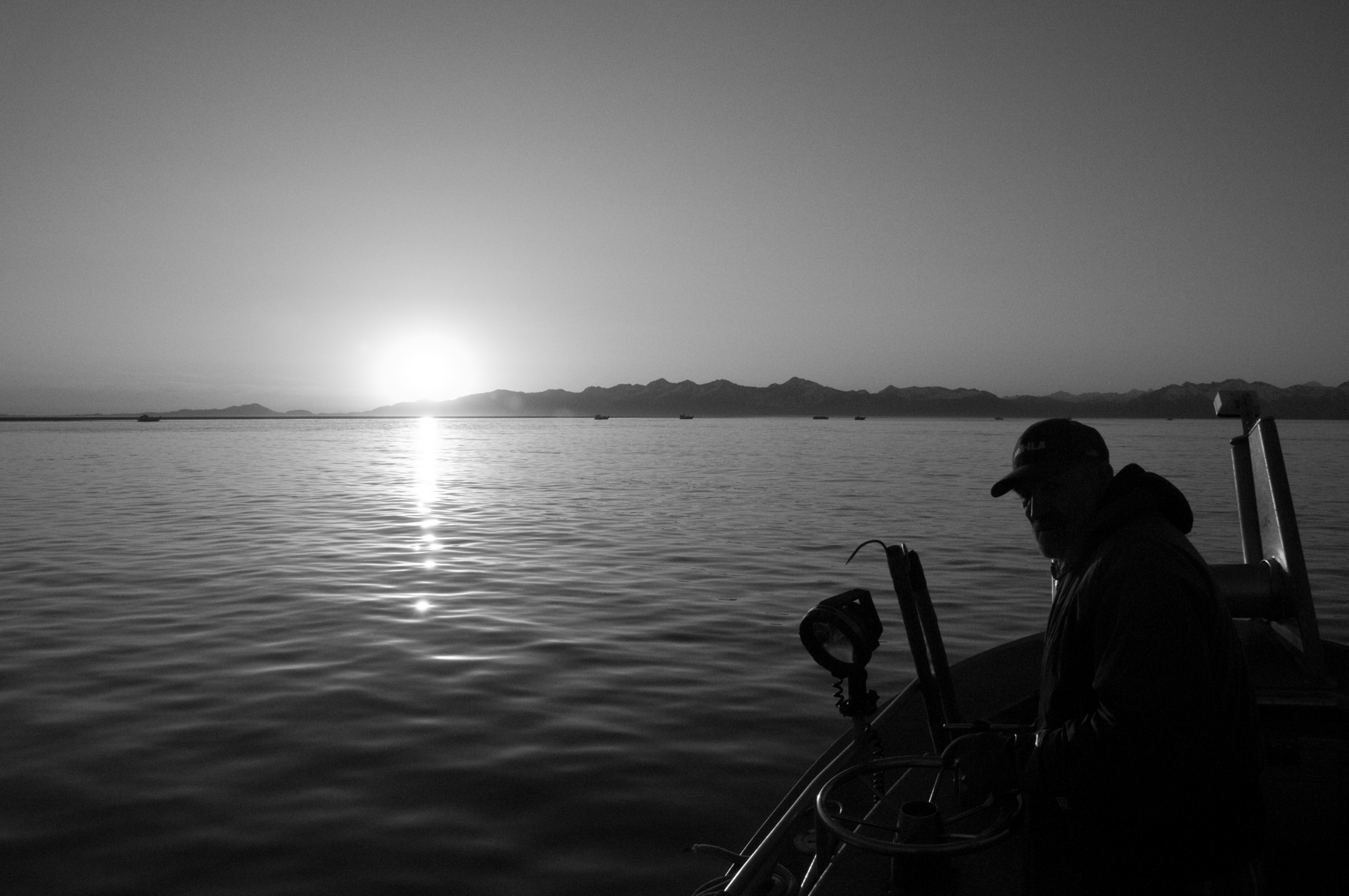 Auto-generated description: A person is standing on a boat on a calm body of water, watching the sun set or rise over distant mountains.