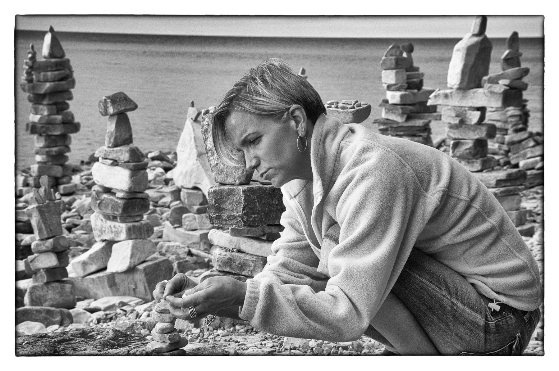Auto-generated description: A person is carefully stacking rocks on a beach surrounded by several balanced rock formations.