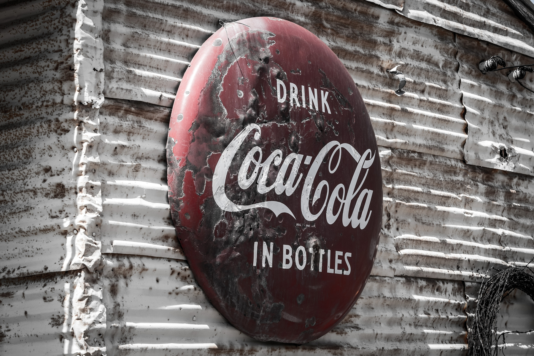 Auto-generated description: A weathered vintage Coca-Cola sign is mounted on a corrugated metal wall.