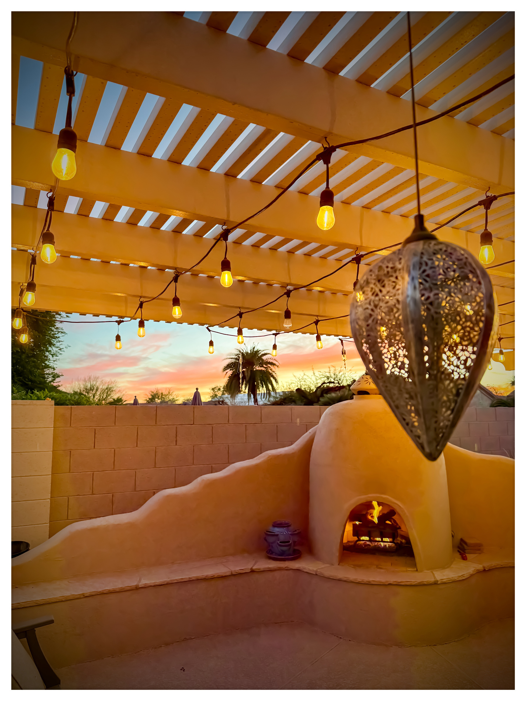 A cozy outdoor patio features a lit fireplace, hanging lights, and a decorative lantern against a colorful sunset sky.