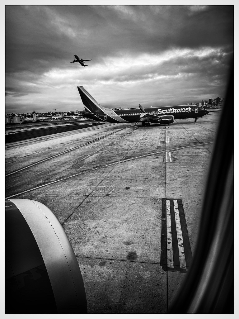 Auto-generated description: A black and white view from an airplane window shows a Southwest Airlines plane on the tarmac and another plane taking off in the cloudy sky.