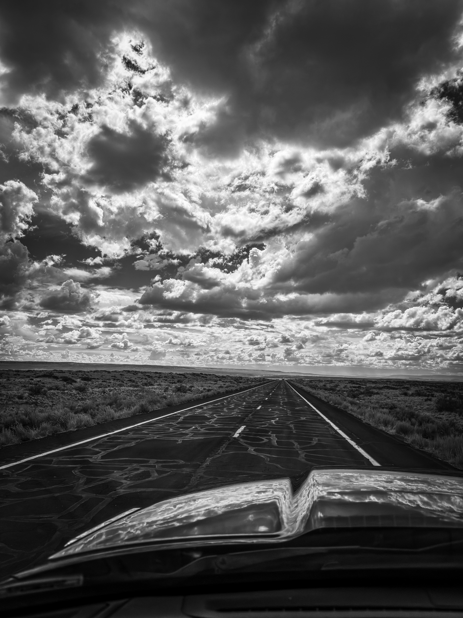 Auto-generated description: A dramatic black-and-white view from a car's windshield shows a long, straight road disappearing into the horizon under a sky filled with dynamic, textured clouds.