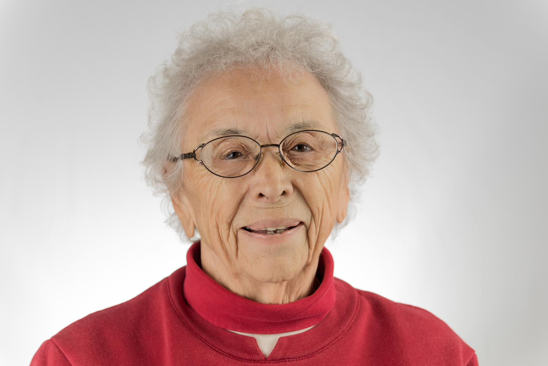 Auto-generated description: An elderly person with short, curly gray hair and glasses is wearing a red top against a neutral background.