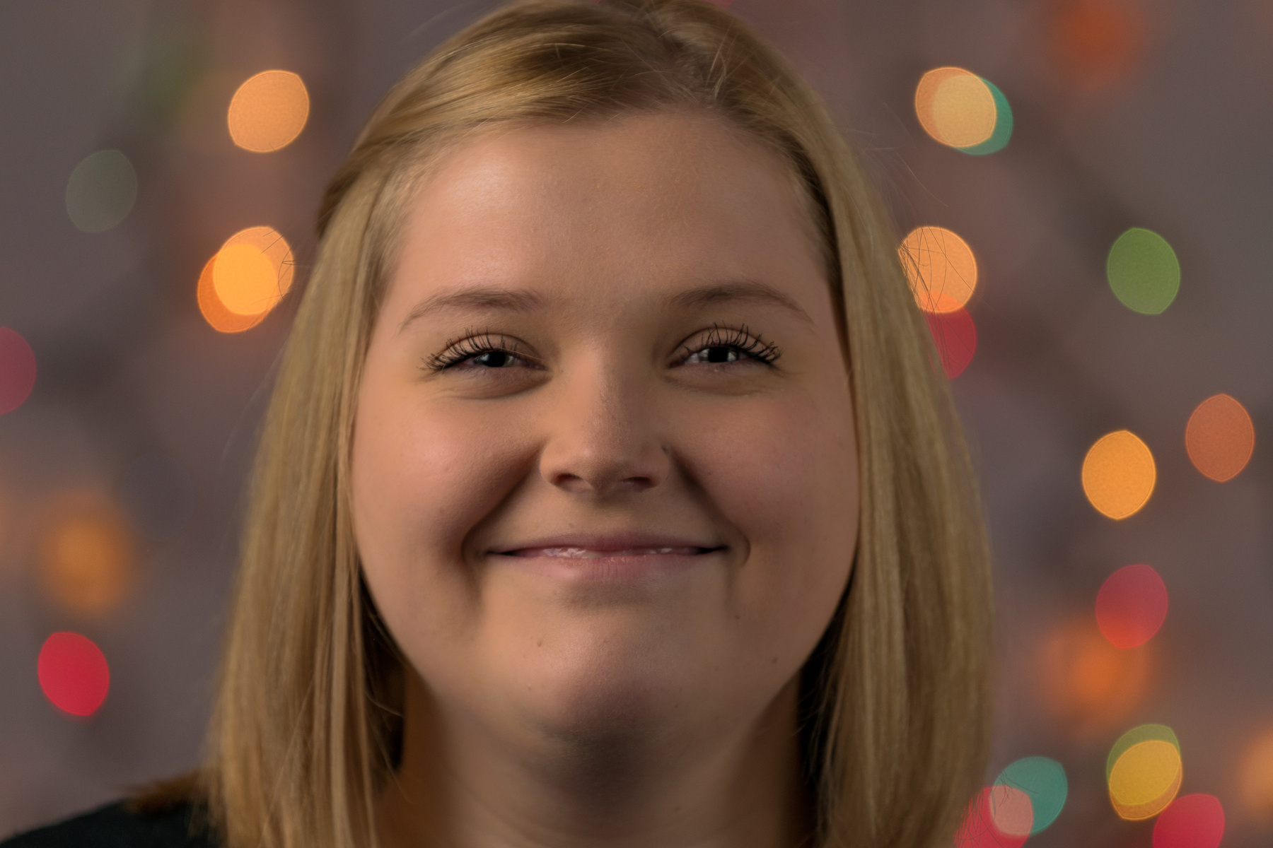 Auto-generated description: A person with blonde hair is smiling in front of a background with colorful, blurred lights.