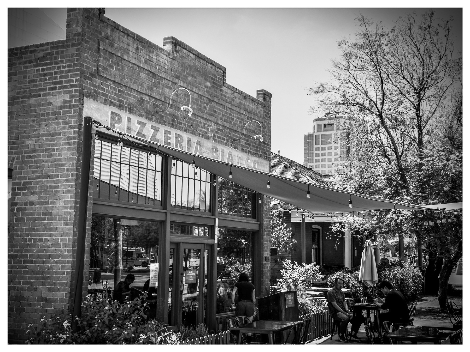 Auto-generated description: A rustic brick building houses a pizzeria with outdoor seating, shaded by hanging canopies and surrounded by trees.