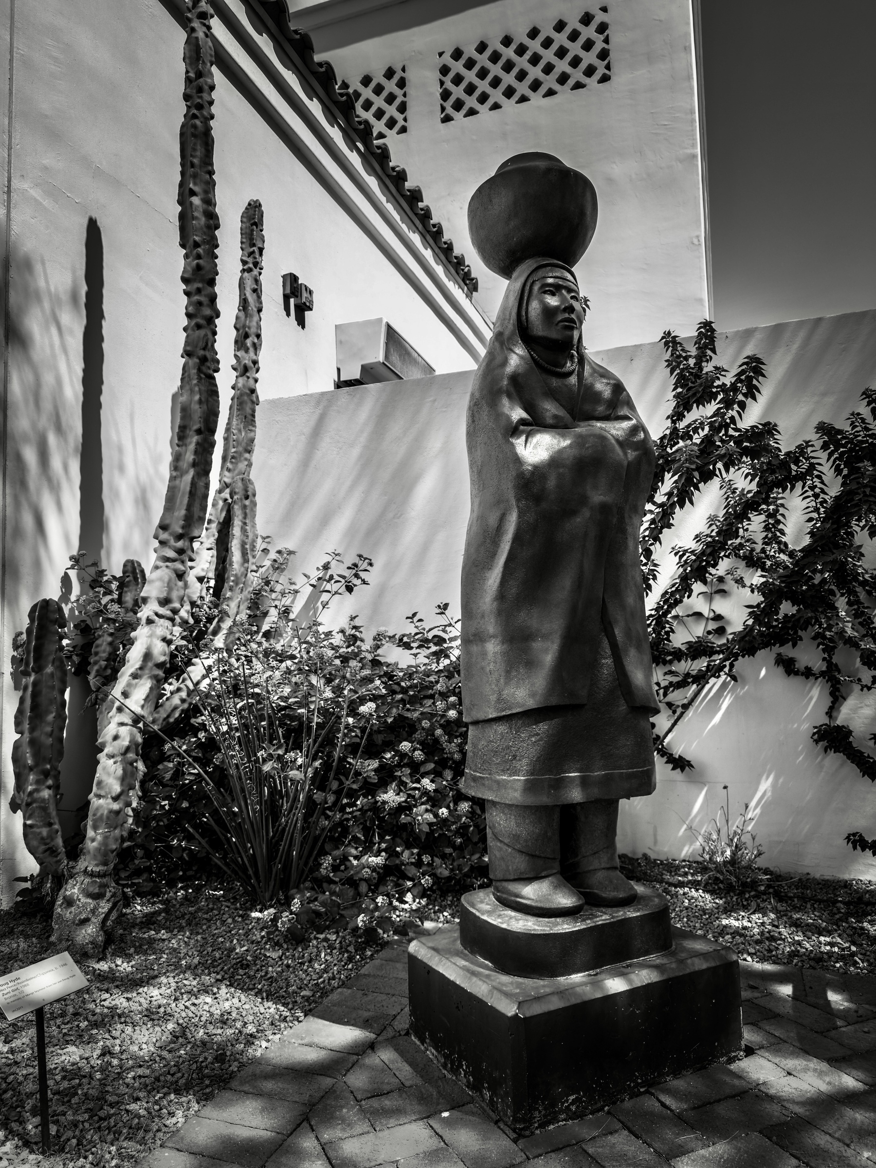 Auto-generated description: A black and white photograph shows a statue of a person holding a large rounded object on their head, surrounded by cactus plants and a building wall.