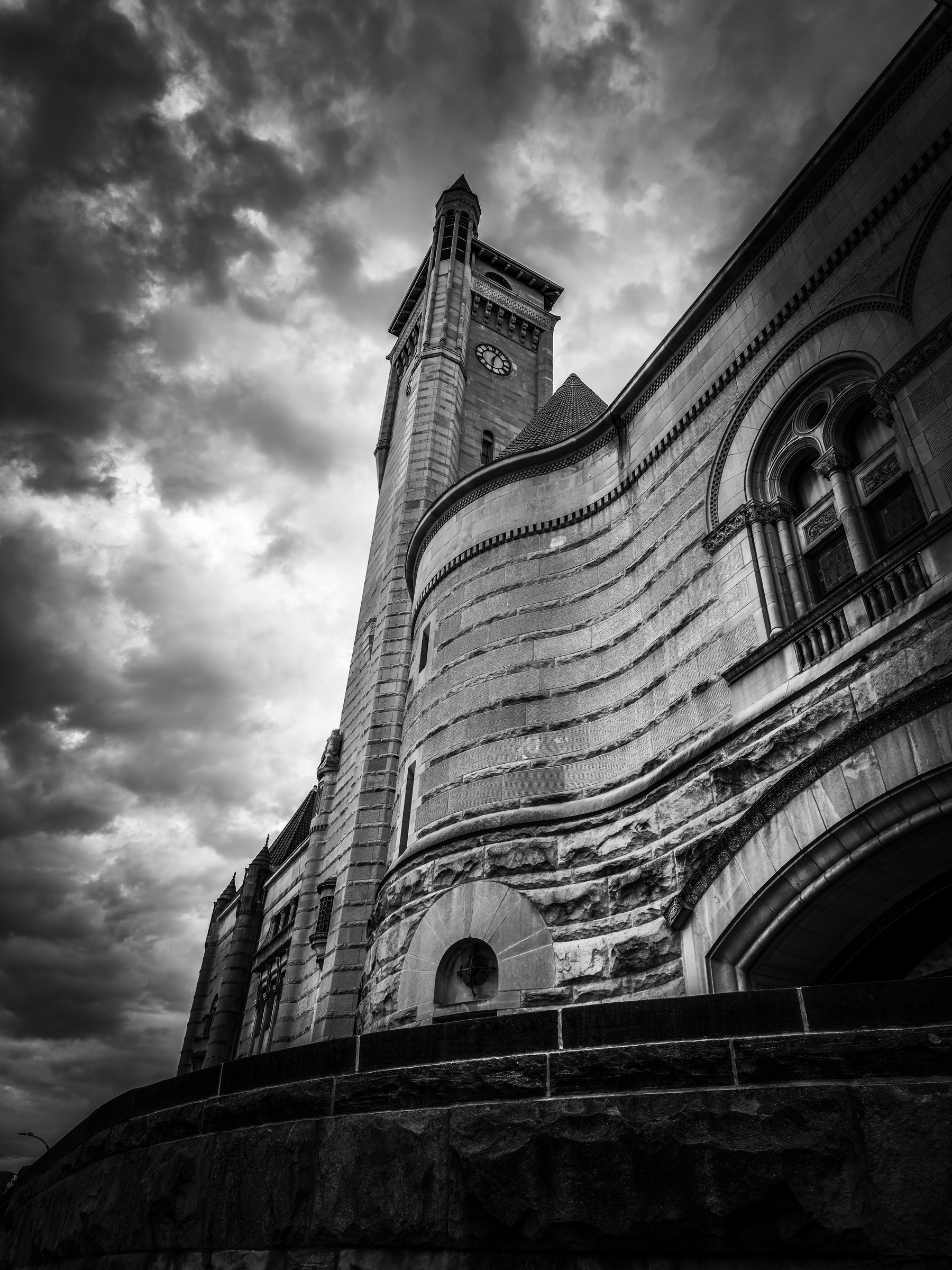 Auto-generated description: A dramatic black and white photograph captures a historic stone building with a tall clock tower under a moody, cloudy sky.