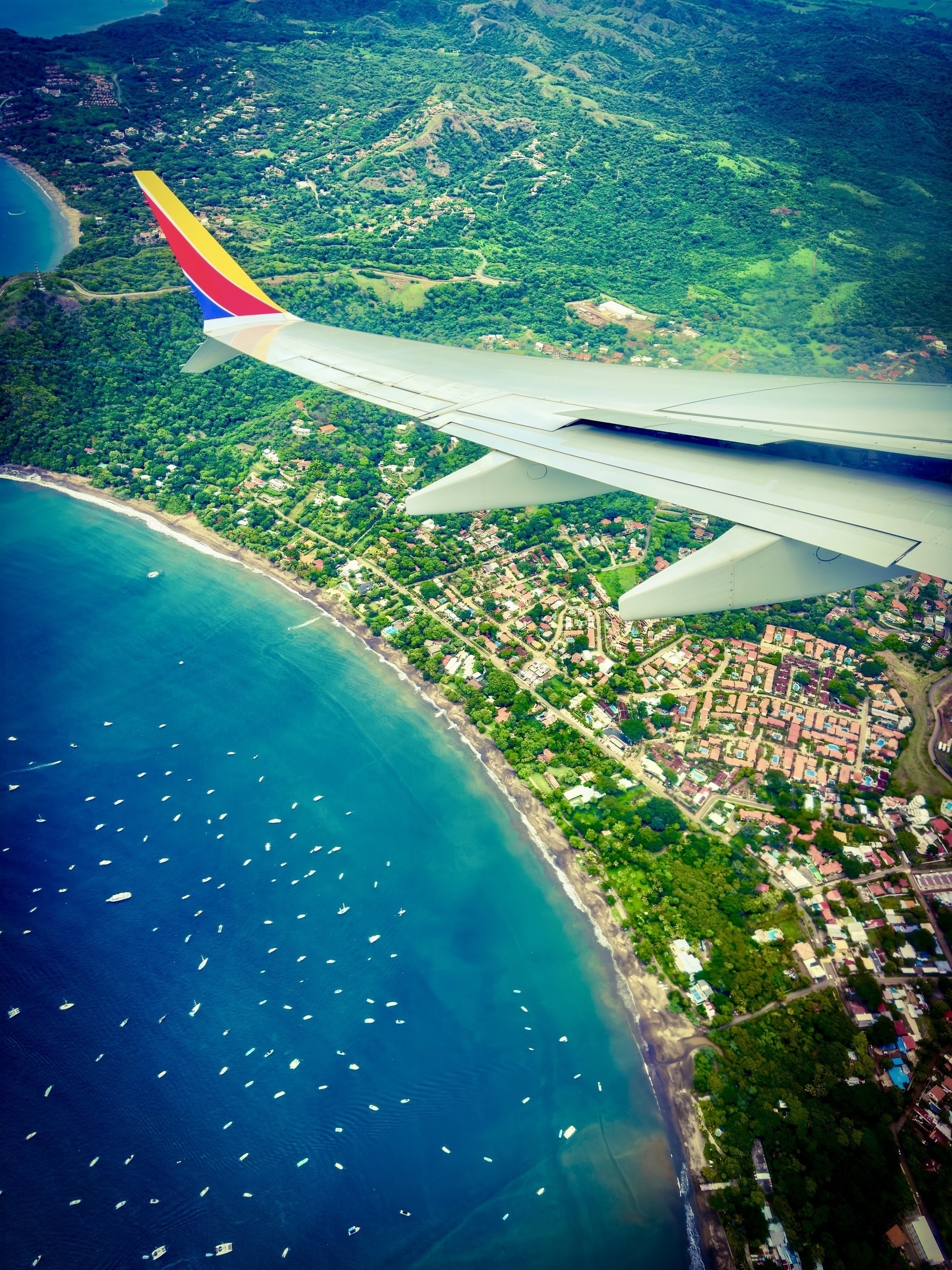 Auto-generated description: An aerial view from an airplane window showcases a coastal landscape with a vibrant turquoise sea, numerous small boats, and lush green land interspersed with buildings.