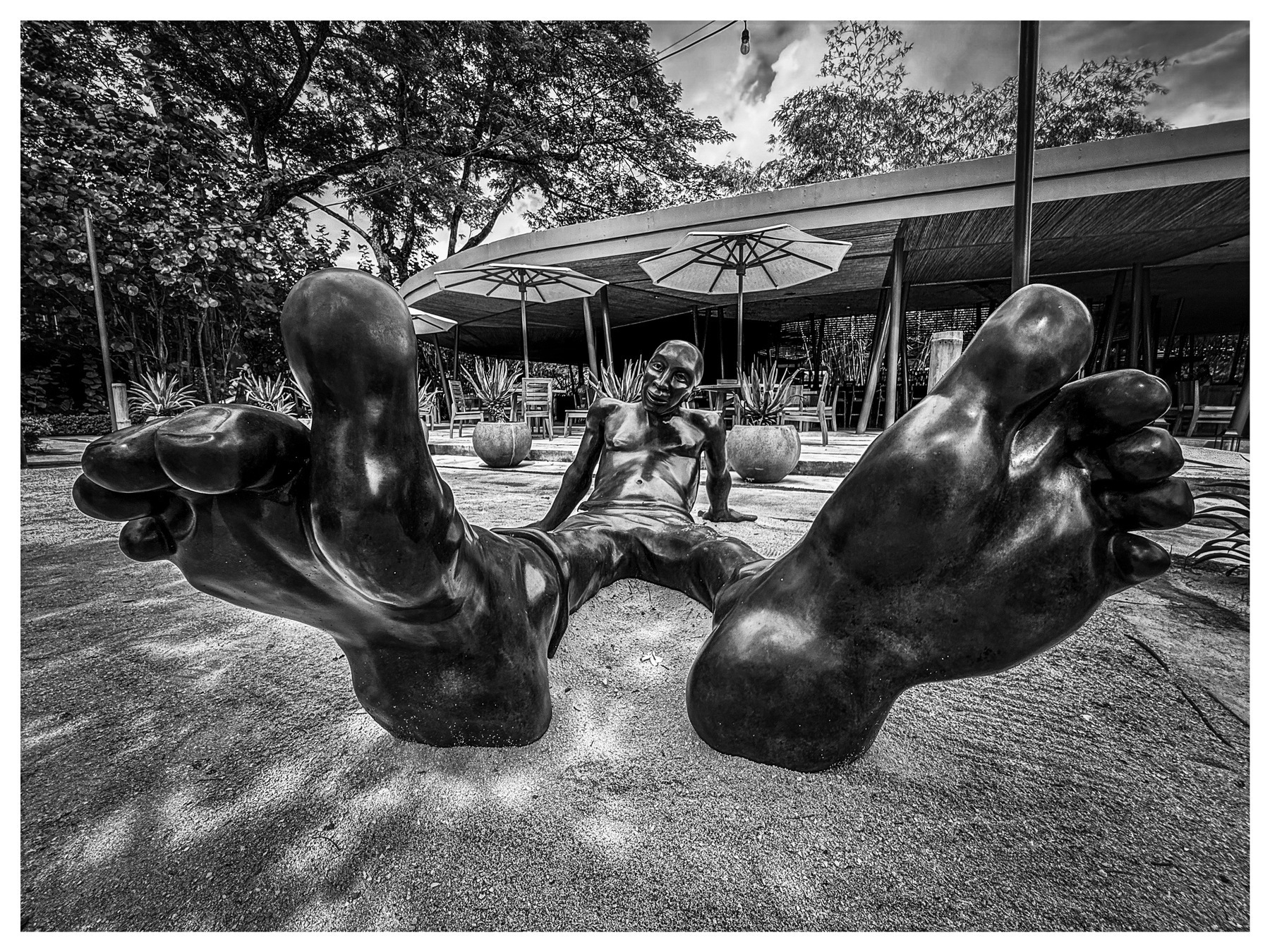 Auto-generated description: A large sculpture of a person with oversized feet lies outdoors near a modern structure with umbrellas and plants.