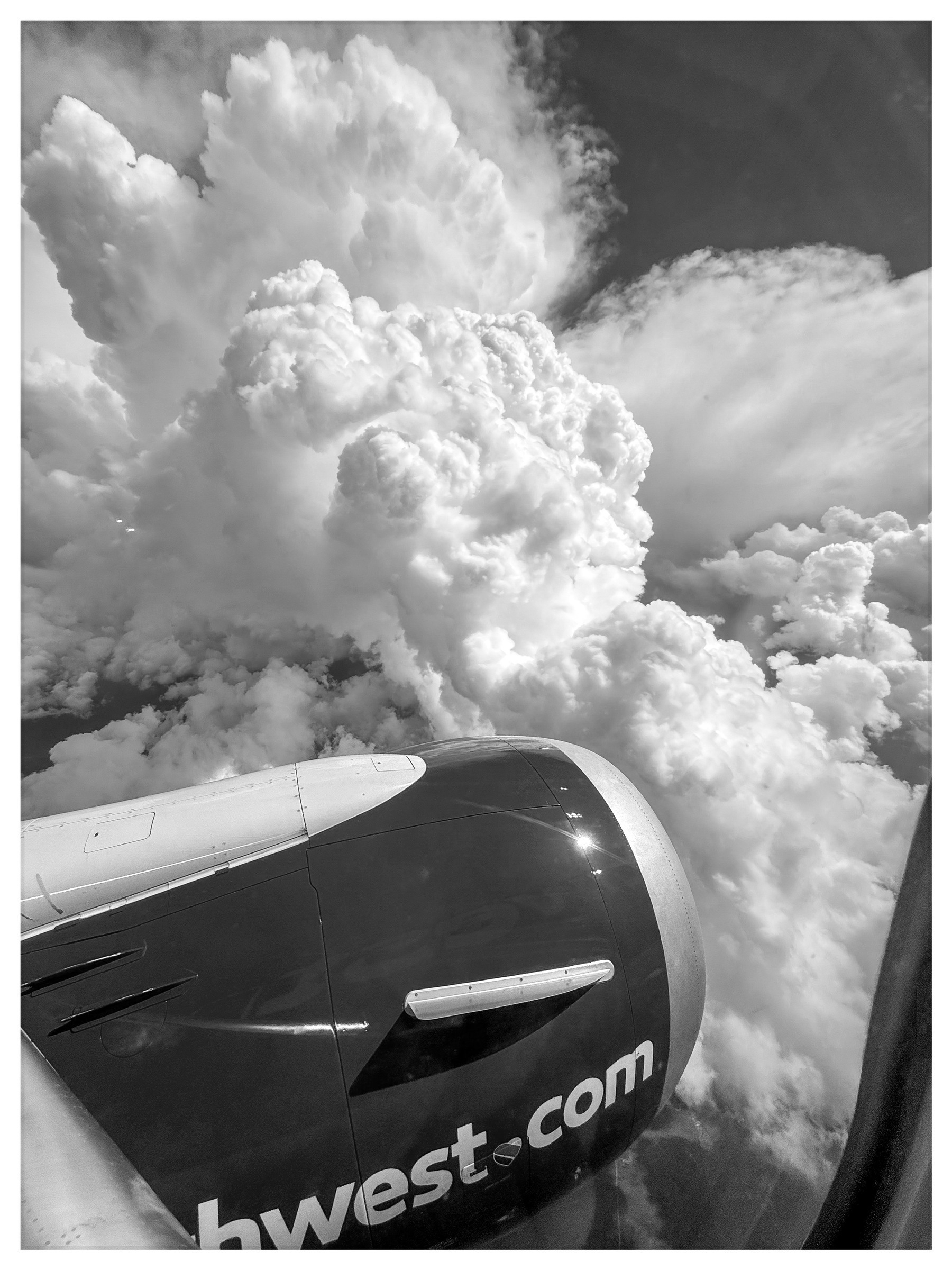 Auto-generated description: A dramatic black and white view from a plane window captures fluffy, towering clouds alongside the engine with part of the plane's exterior visible.