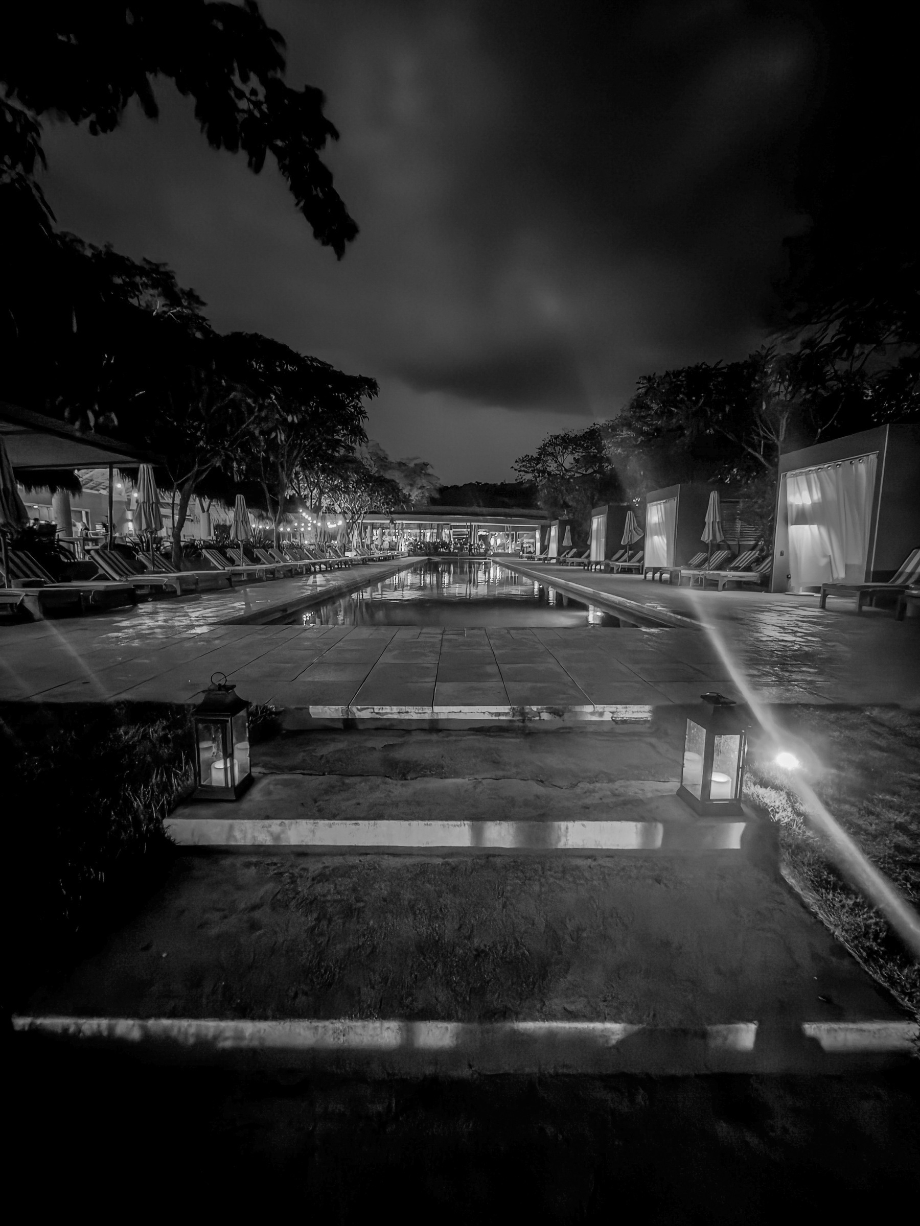 Auto-generated description: A serene nighttime scene features a pool lined with lounge chairs and lanterns, with trees silhouetted against a cloudy sky.