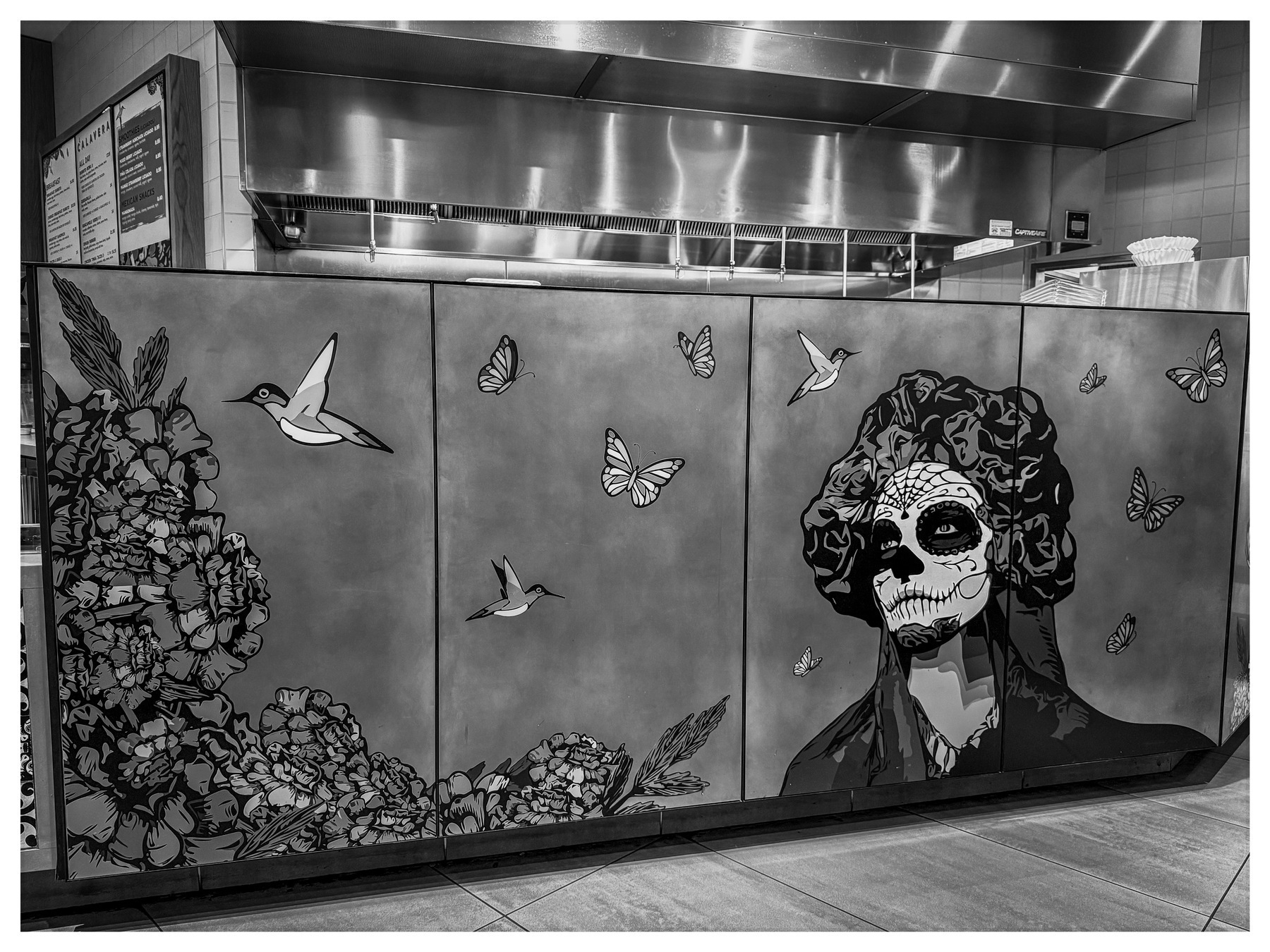 Auto-generated description: A mural depicting a skeletal figure surrounded by hummingbirds, butterflies, and floral motifs is displayed on the front of a counter in a stainless steel kitchen setting.