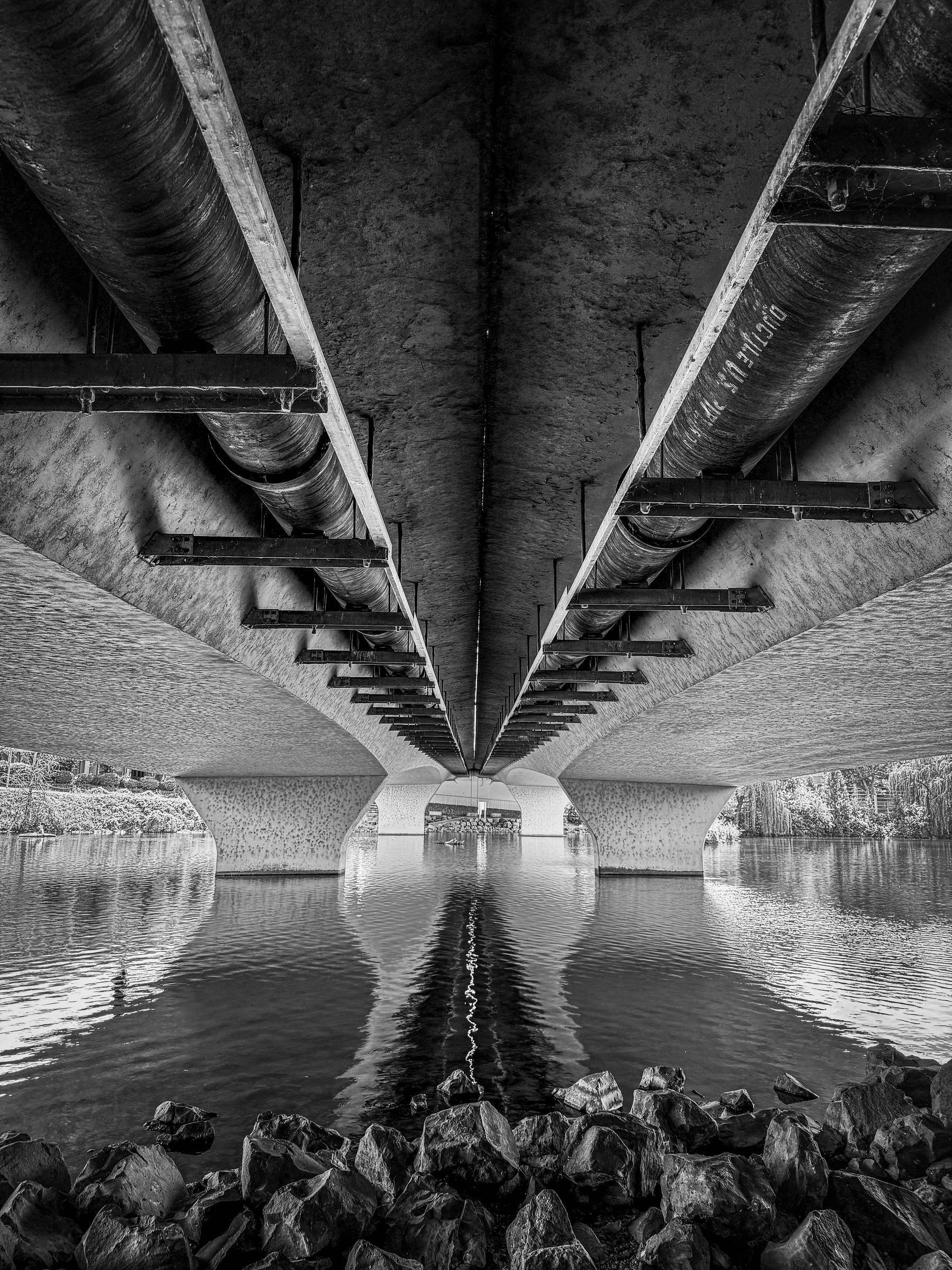 Auto-generated description: A symmetrical black and white view of the underside of a bridge with reflections on a calm body of water and rocks in the foreground.
