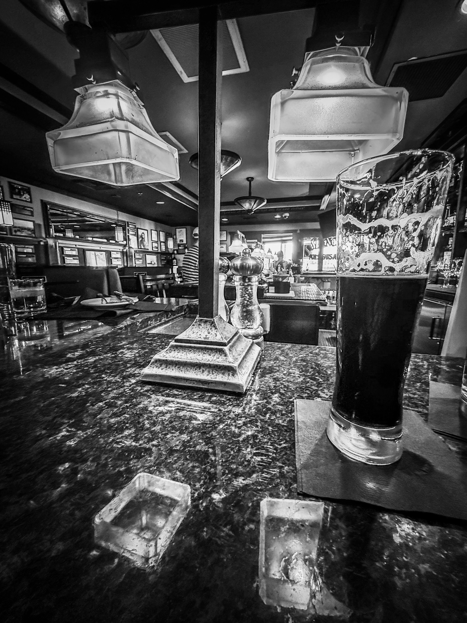 Auto-generated description: A black-and-white photo of a dimly lit bar features a full pint of dark beer on a reflective counter and overhead pendant lights.