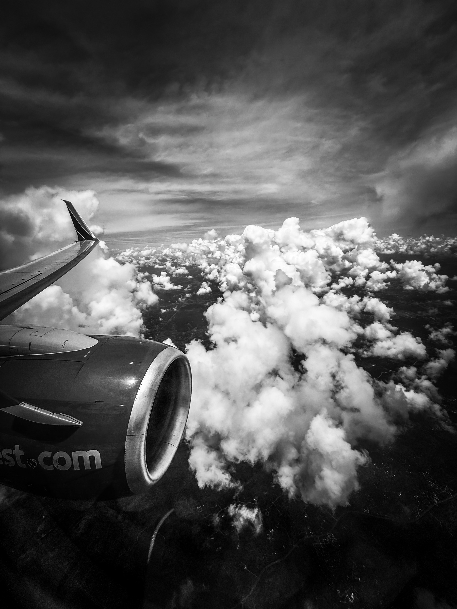 Auto-generated description: A black and white aerial view from an airplane window shows dramatic clouds and the plane's engine against a backdrop of vast skies.
