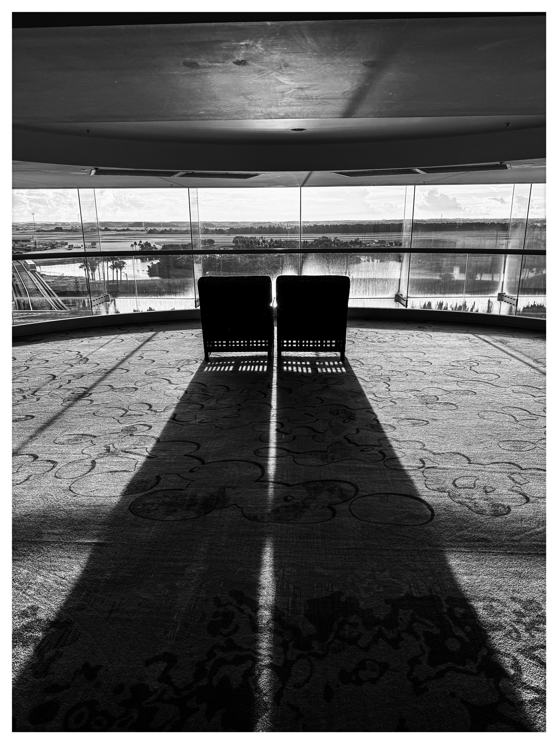 Auto-generated description: Two chairs cast long shadows on a patterned carpeted floor in front of a large window with a view of a river and landscape.