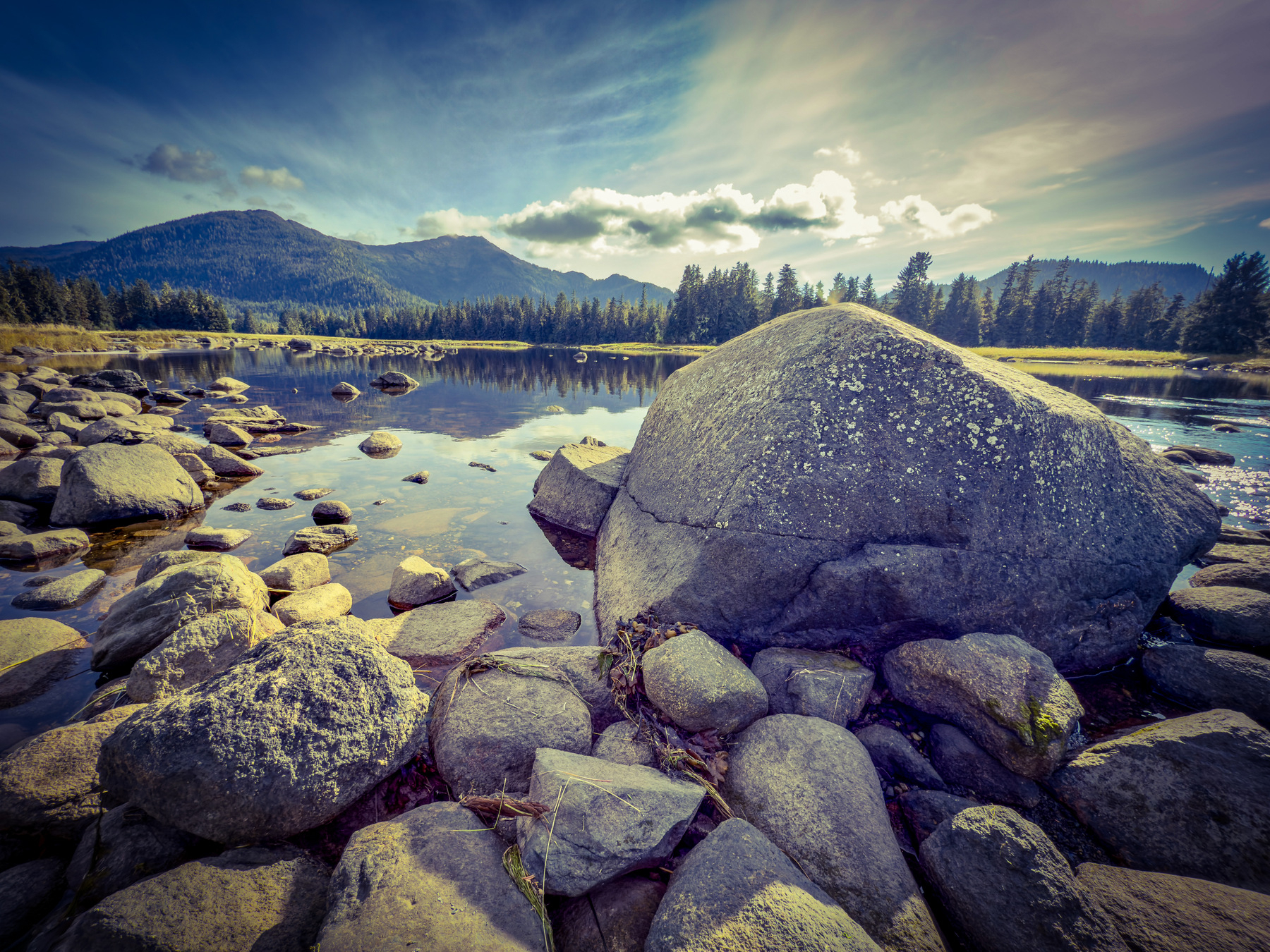 Auto-generated description: A serene mountain landscape features a rocky shoreline bordering a reflective lake under a partly cloudy sky.