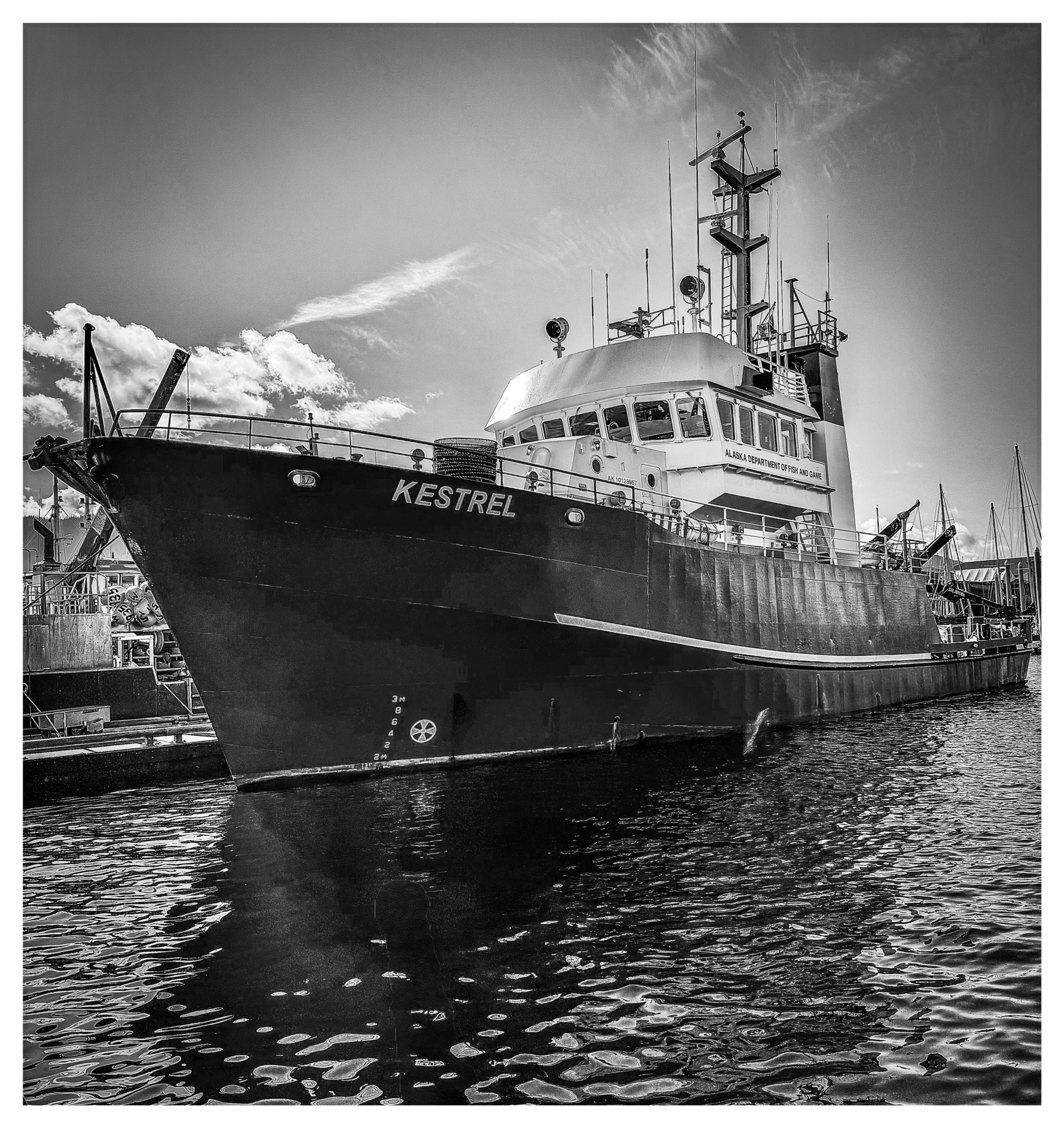 Auto-generated description: A large ship named Kestrel is docked at a harbor under a partly cloudy sky, captured in black and white.