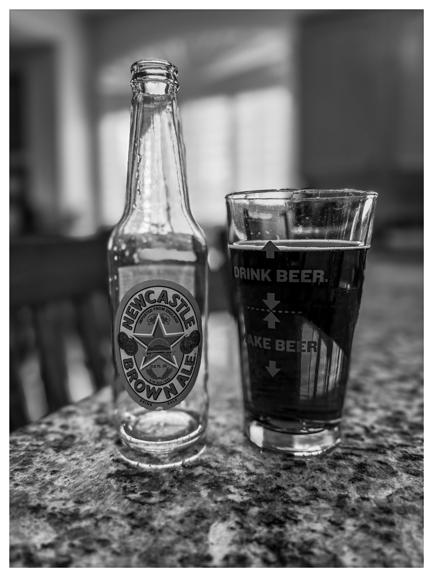 Auto-generated description: A bottle of Newcastle Brown Ale sits next to a filled pint glass with humorous text, both on a granite countertop.