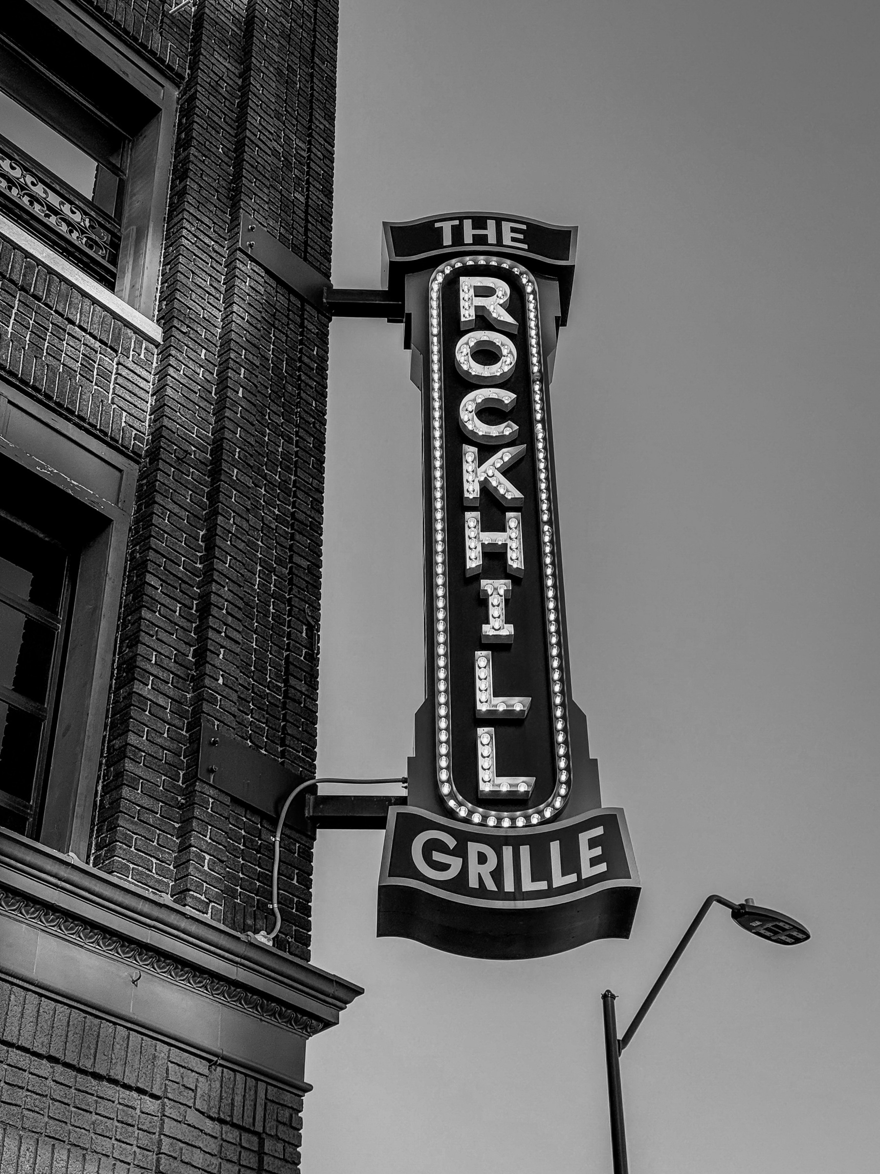 Auto-generated description: A tall vertical sign attached to a brick building reads The Rockhill Grille.