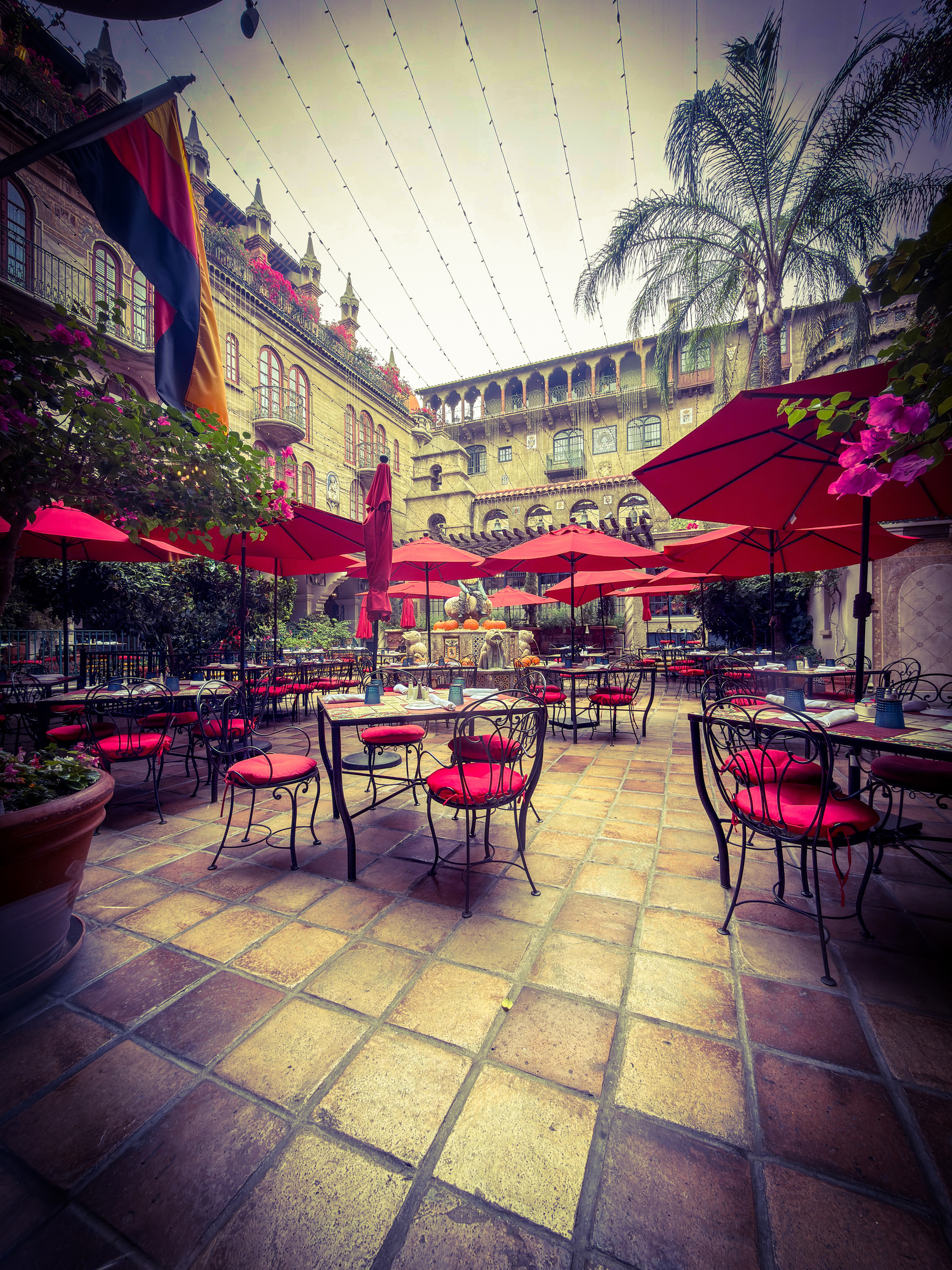 Auto-generated description: A charming courtyard features tiled flooring, red umbrellas, iron chairs with red cushions, and ornate architecture in the background.