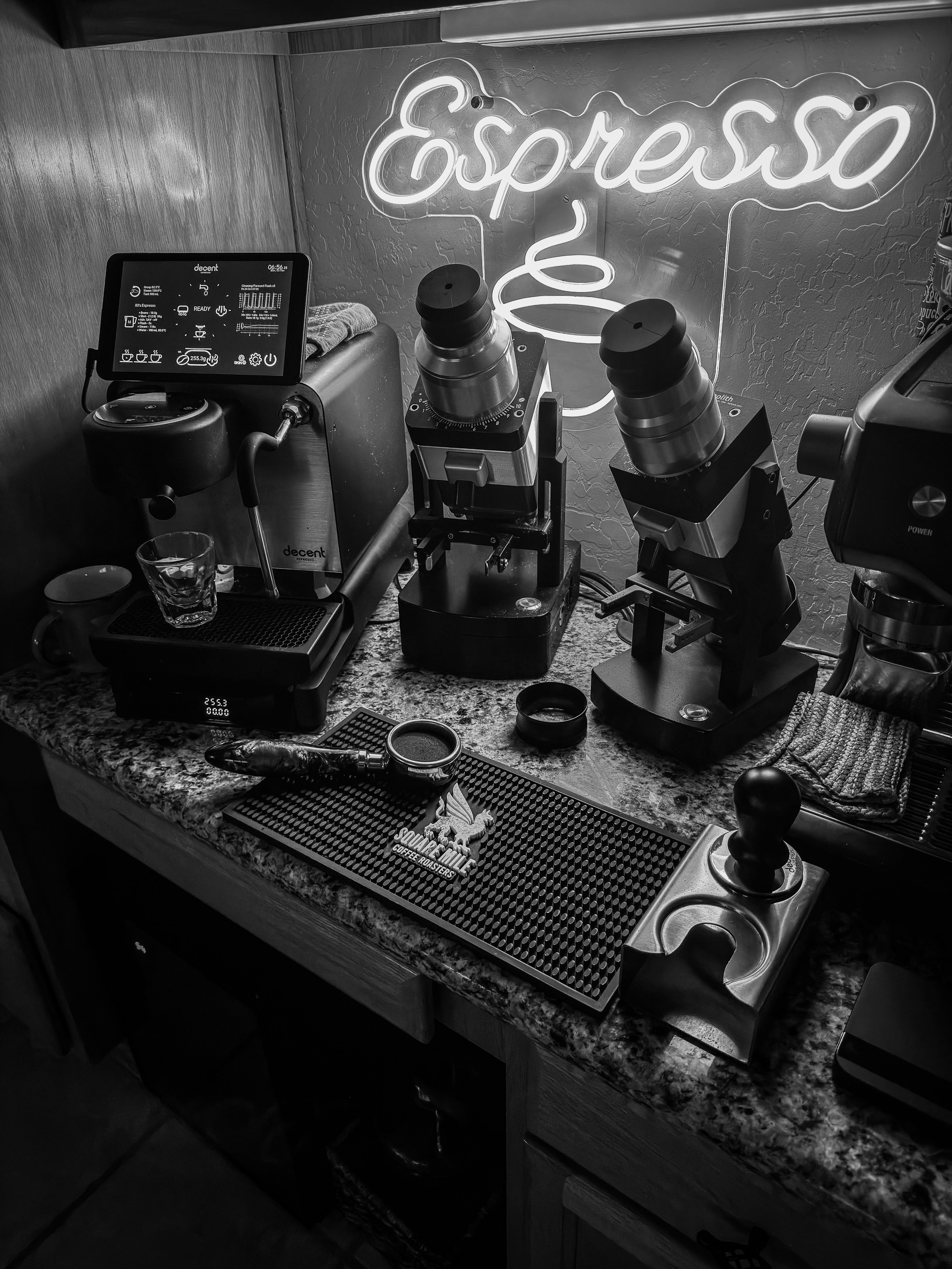 Auto-generated description: A black and white photo featuring a coffee setup with espresso machines, a digital coffee scale, a tamper, and a neon sign reading Espresso.