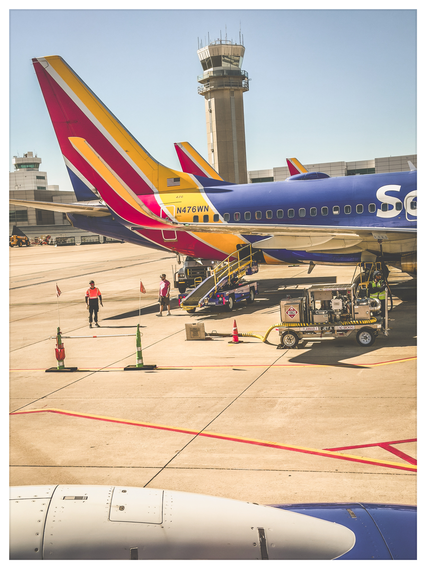 Auto-generated description: An airplane is parked at an airport gate with workers and ground support equipment around, and a control tower in the background.