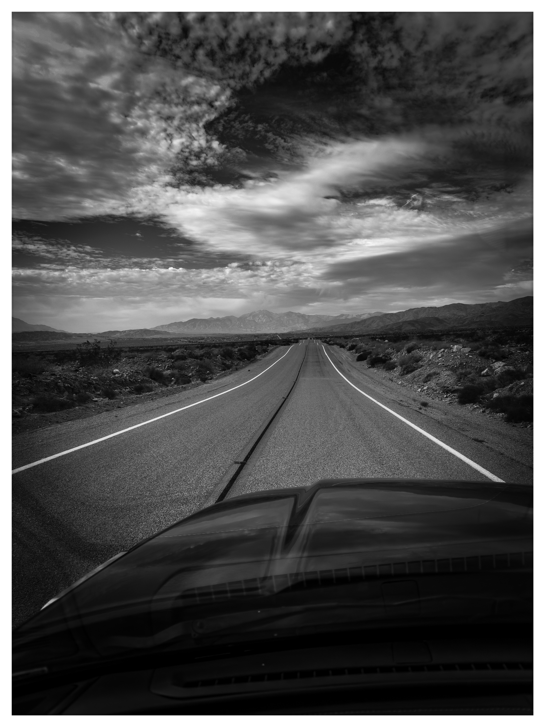 Auto-generated description: A long, empty road stretches into the distance under a dramatic, cloudy sky, as seen from the perspective of a vehicle.
