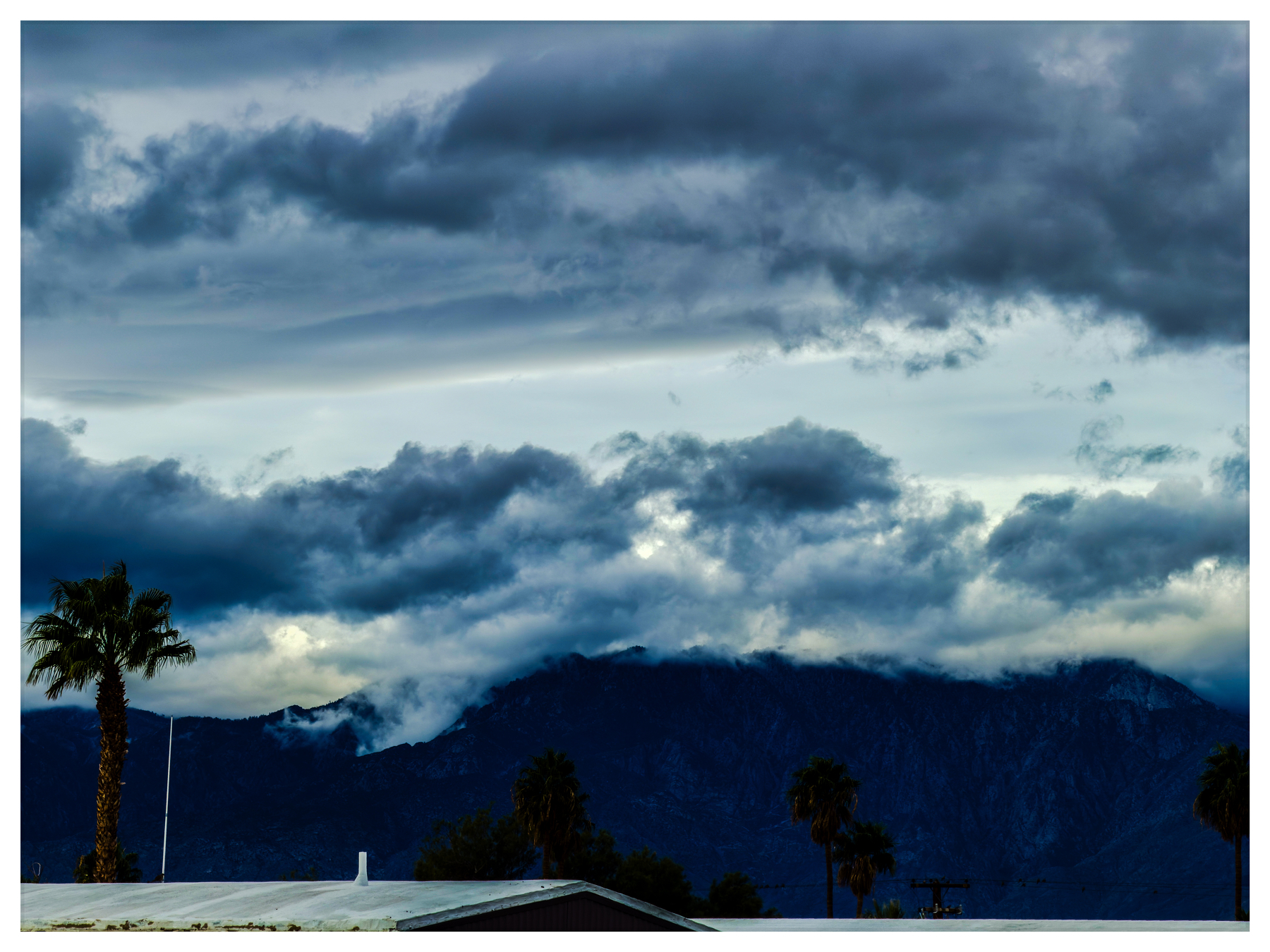 Auto-generated description: Dark, dramatic clouds hover over a mountainous landscape with silhouetted palm trees in the foreground.