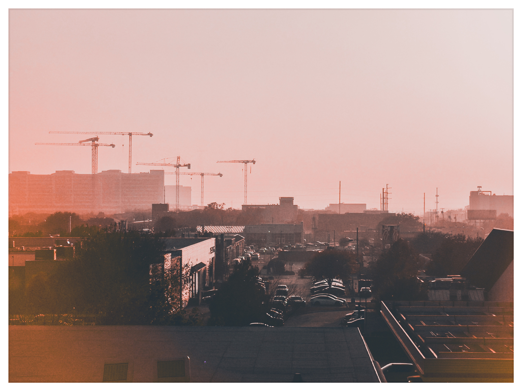 Auto-generated description: A hazy urban landscape features several construction cranes and buildings under a reddish sky.