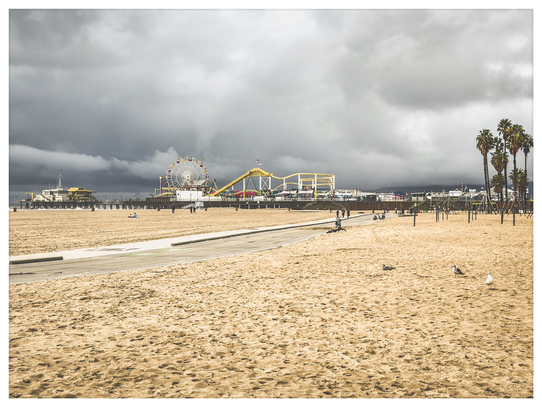Auto-generated description: A sandy beach leads to a vibrant amusement park with a Ferris wheel under a cloudy sky.