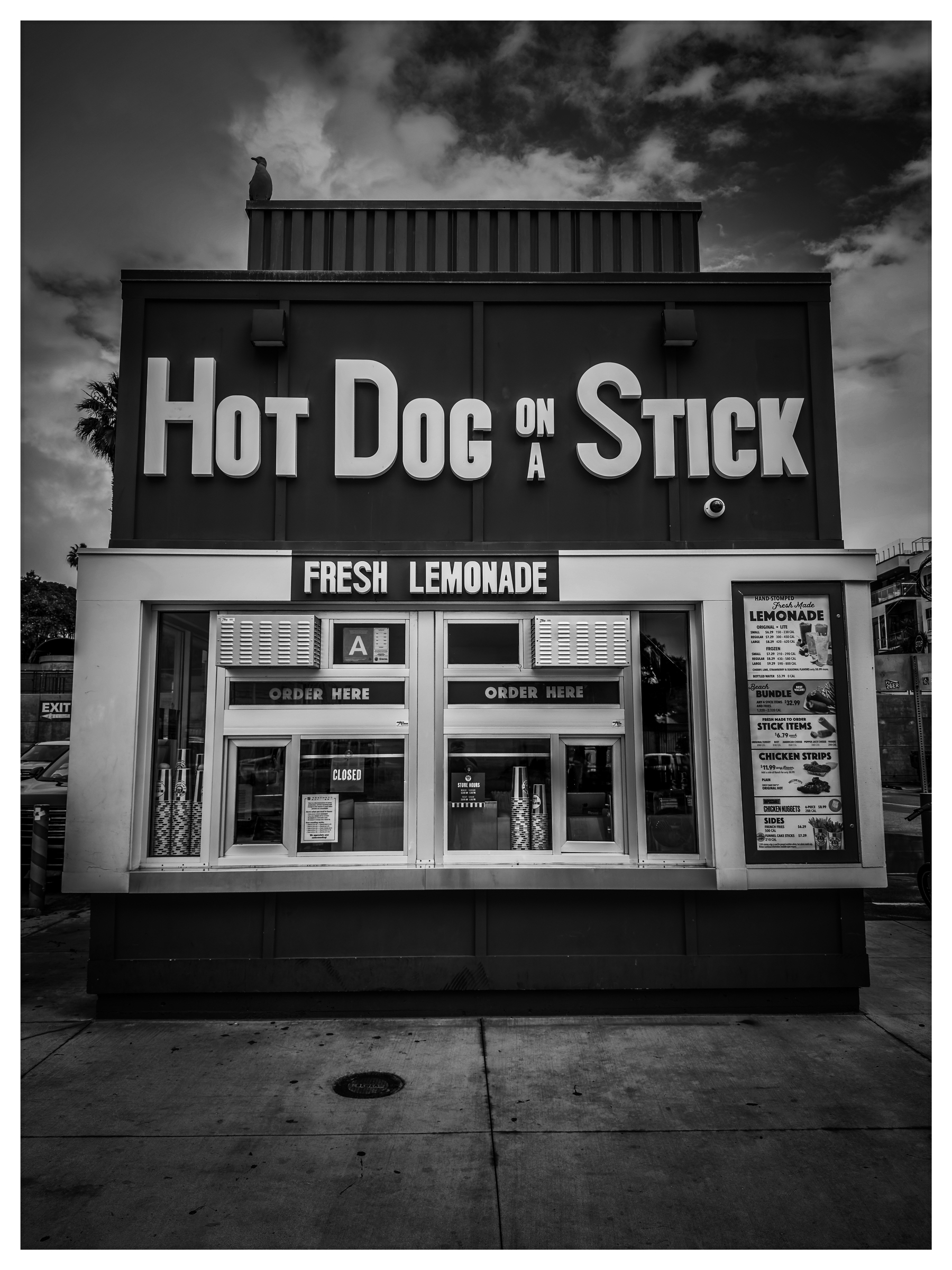 Auto-generated description: A black-and-white photo of a Hot Dog on a Stick stand with prominent signage and menus visible.