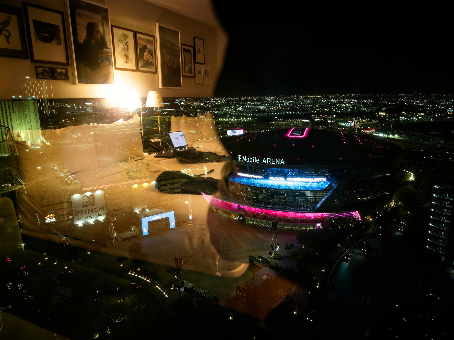 Auto-generated description: A nighttime cityscape is seen through a window, reflecting an interior with framed pictures and a T-Mobile Arena sign illuminated in pink.