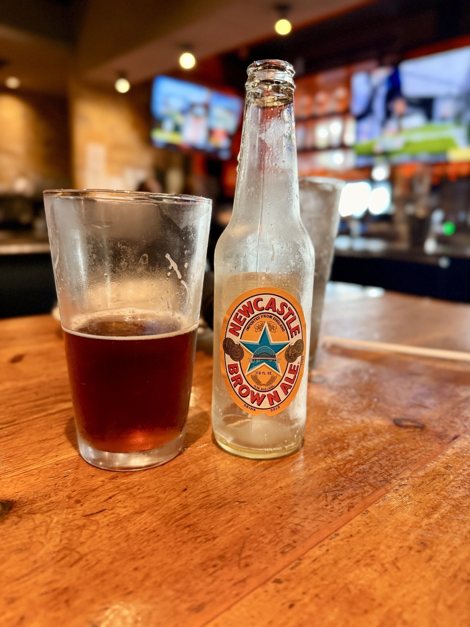 Auto-generated description: A half-full glass of dark beer sits next to an empty Newcastle Brown Ale bottle on a wooden table in a bar.
