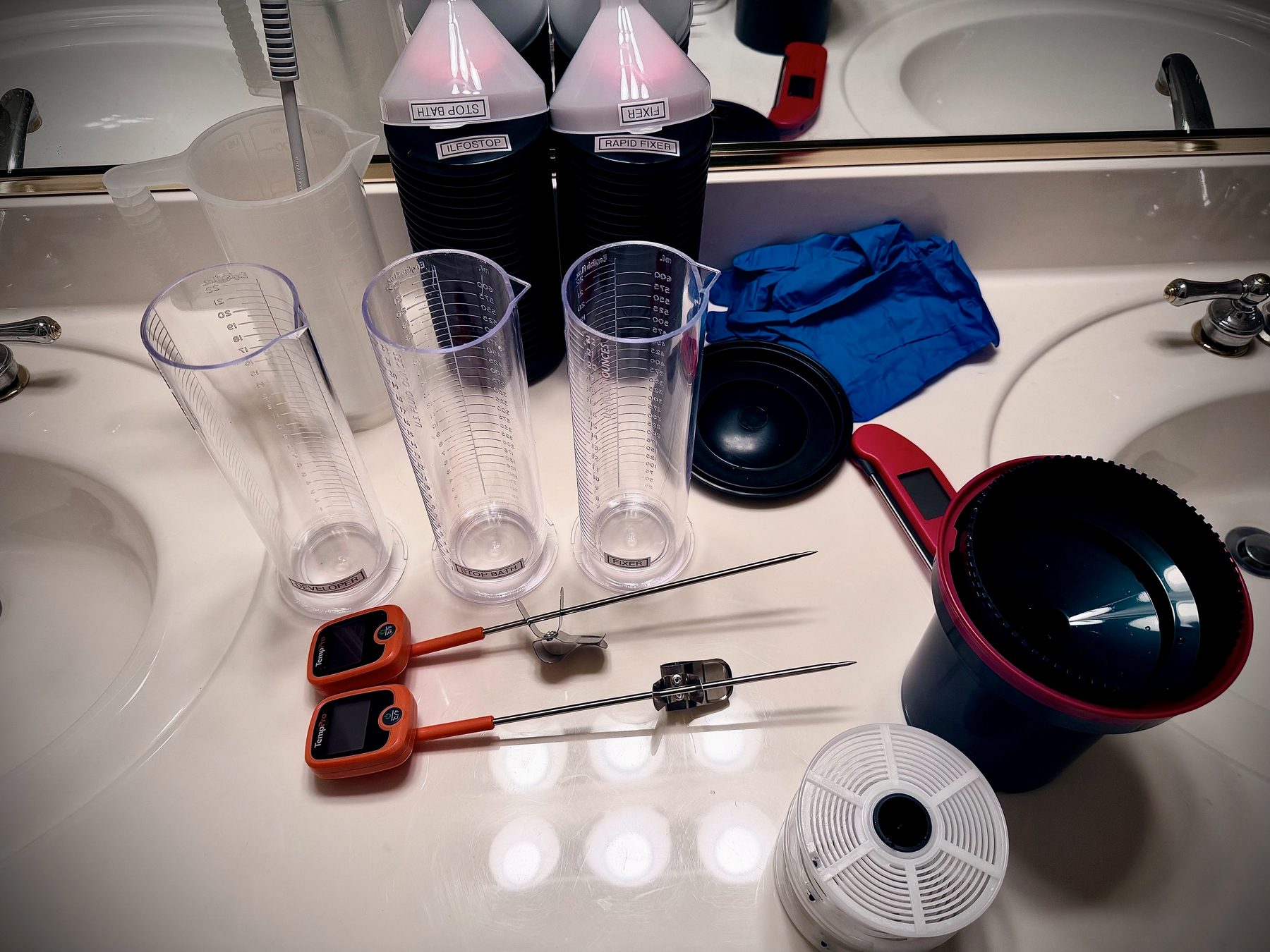 Auto-generated description: Various developing equipment and chemicals are arranged on a bathroom sink countertop, including measuring cups, film developing tanks, thermometers, gloves, and bottles of chemical solutions.