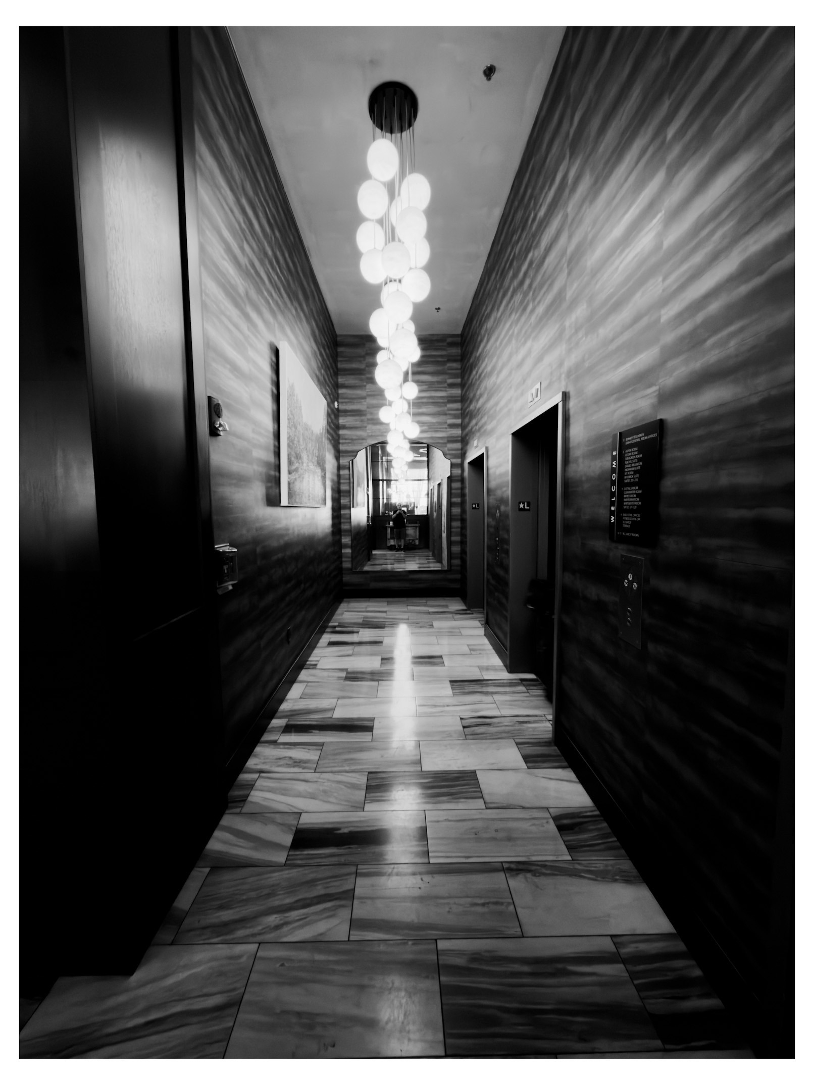 Auto-generated description: A long, narrow hallway with patterned tile flooring, dark textured walls, hanging round lights, and a mirror at the end.