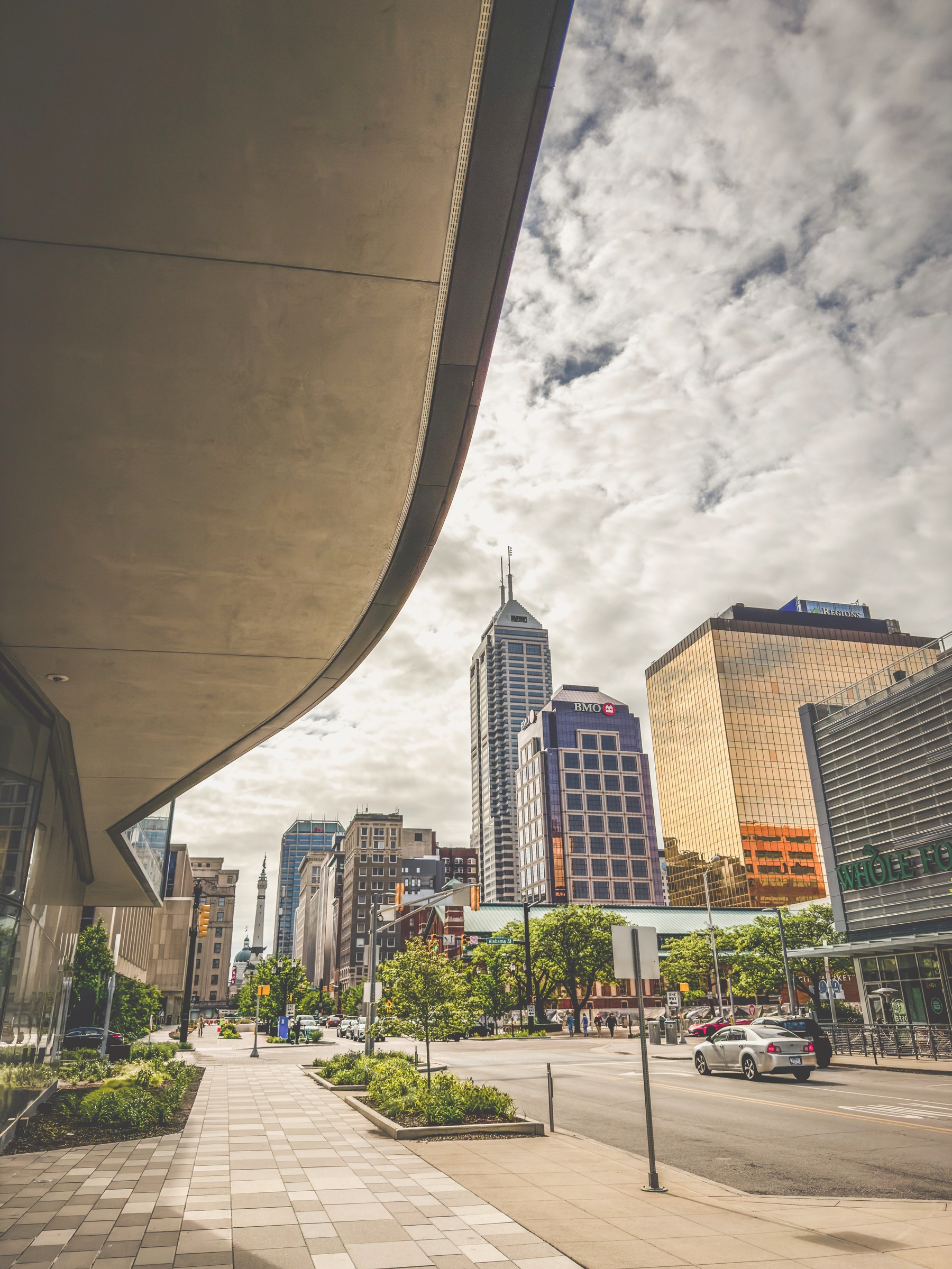 Auto-generated description: A city street scene features modern architecture, towering buildings, and a partly cloudy sky.