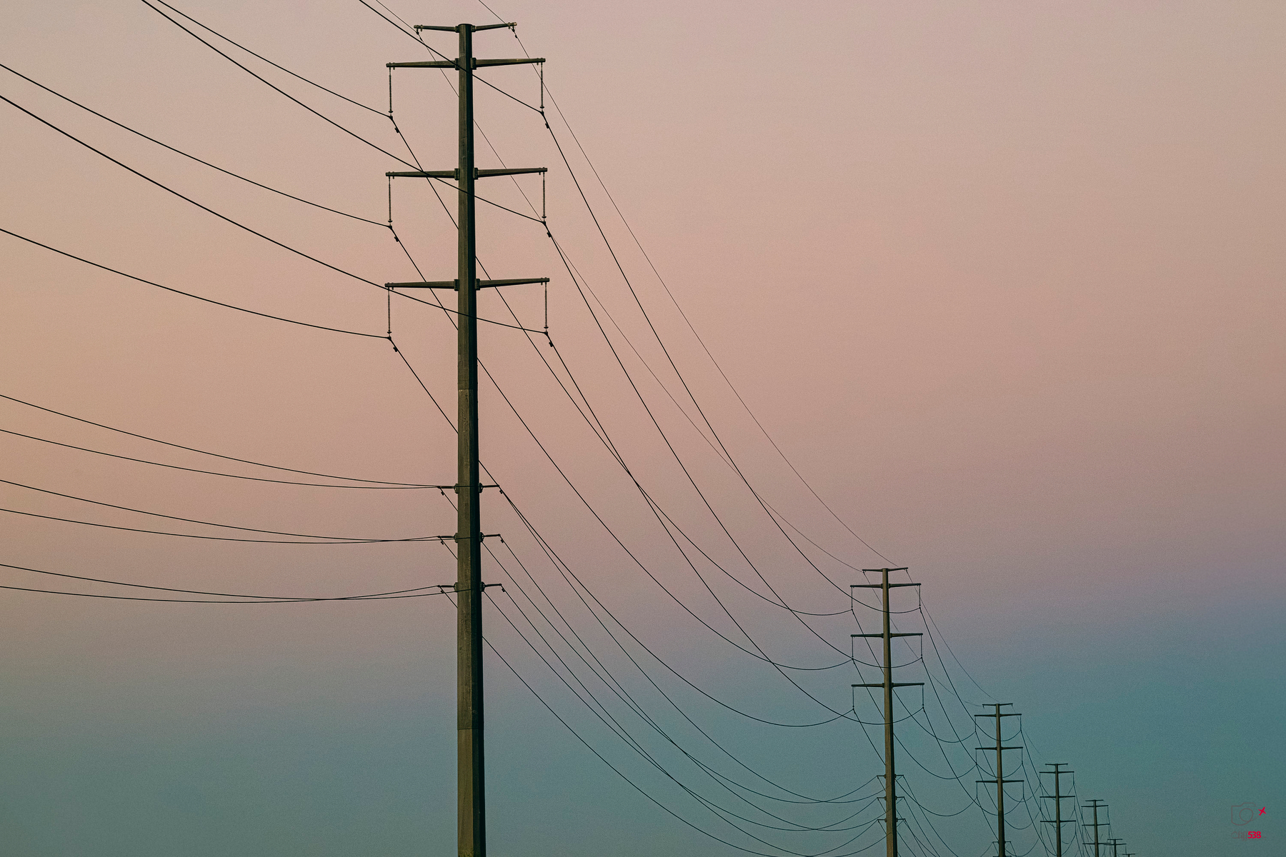 Auto-generated description: A series of power lines and utility poles stretch into the distance against a pastel-colored sky.