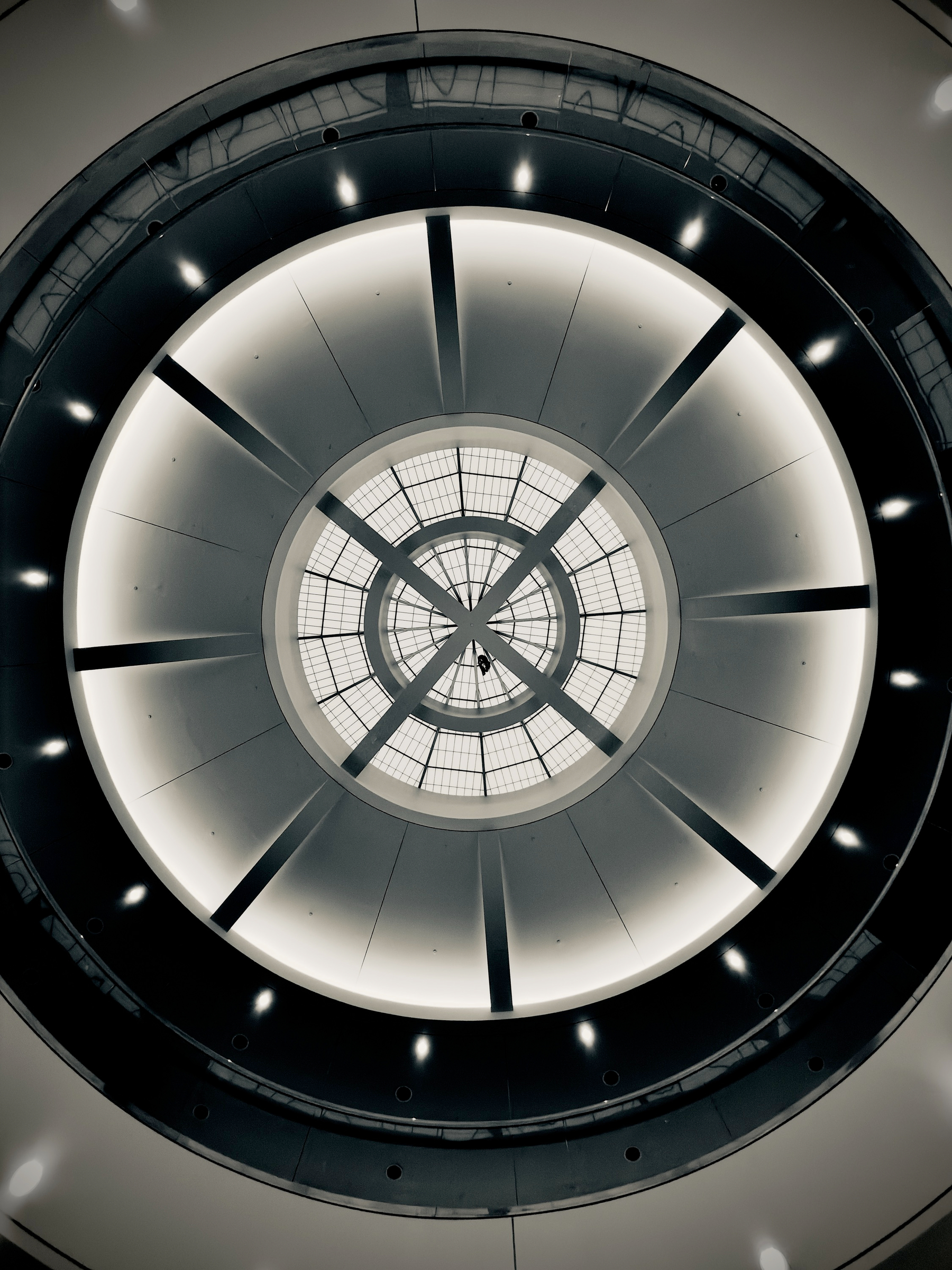 Auto-generated description: A circular, futuristic ceiling design with radiating beams and a central skylight is illuminated by surrounding lights.