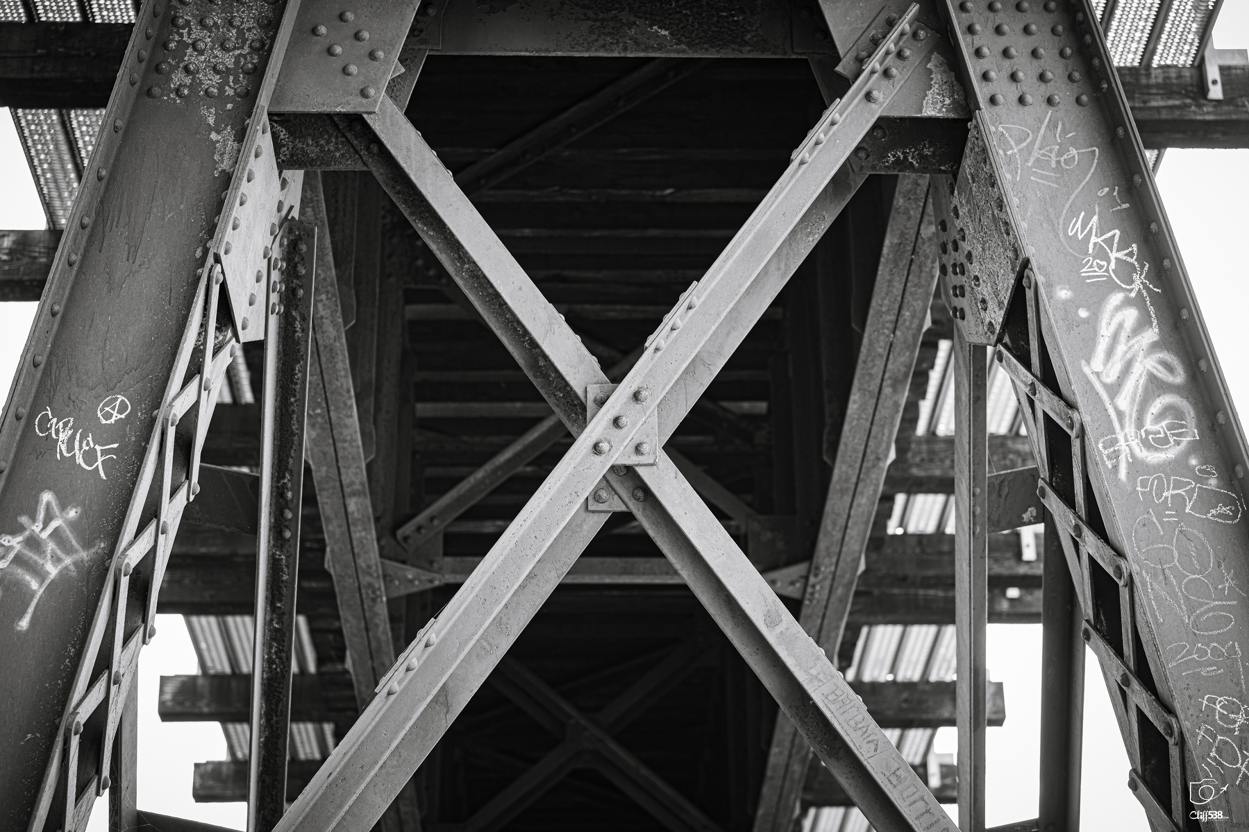 Auto-generated description: A black and white photo captures the underside of a metal bridge with graffiti on its support beams and riveted cross braces.