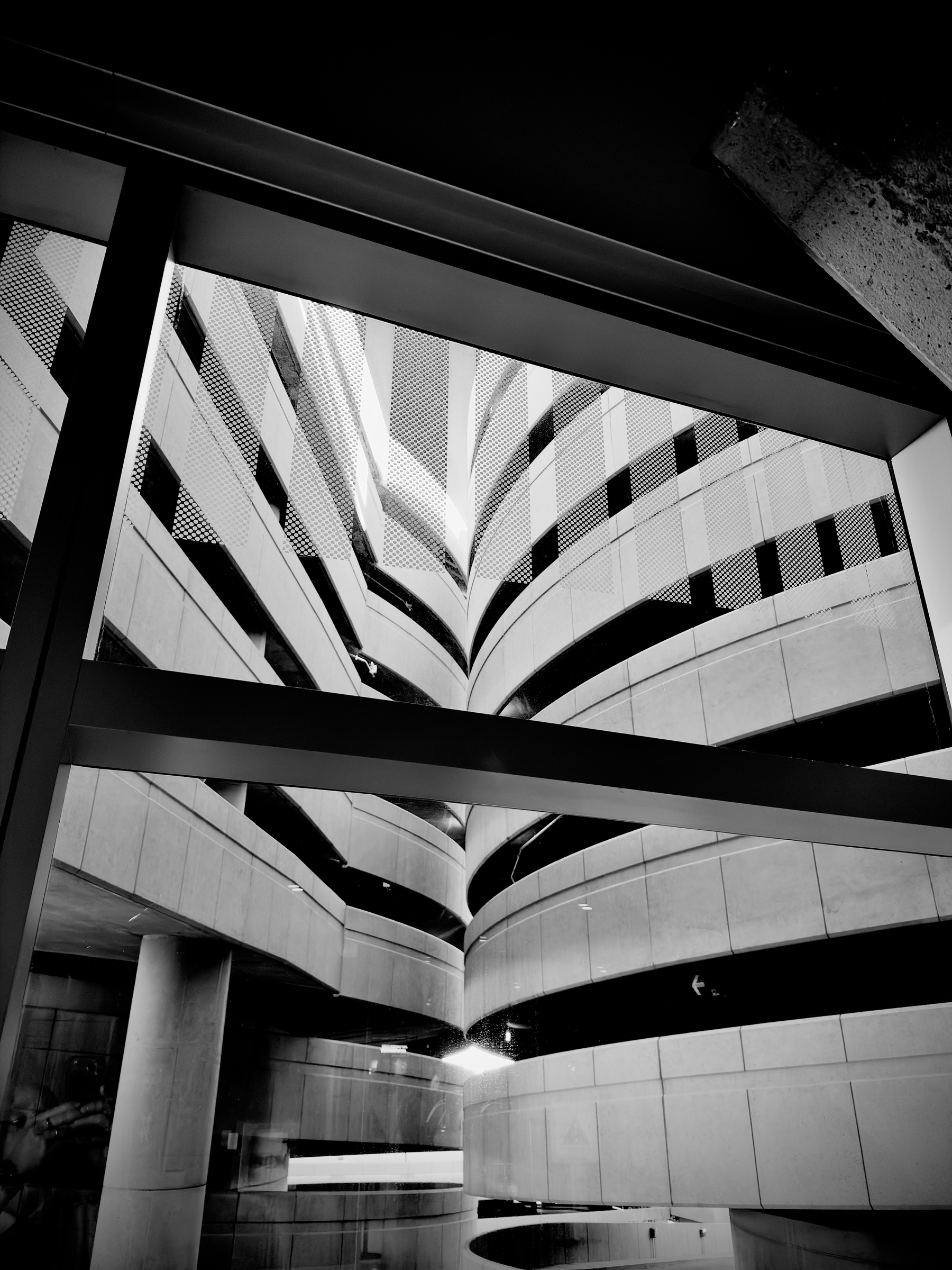 Auto-generated description: A black and white photo capturing the architectural curves and lines of a multi-level parking structure viewed through a window frame.
