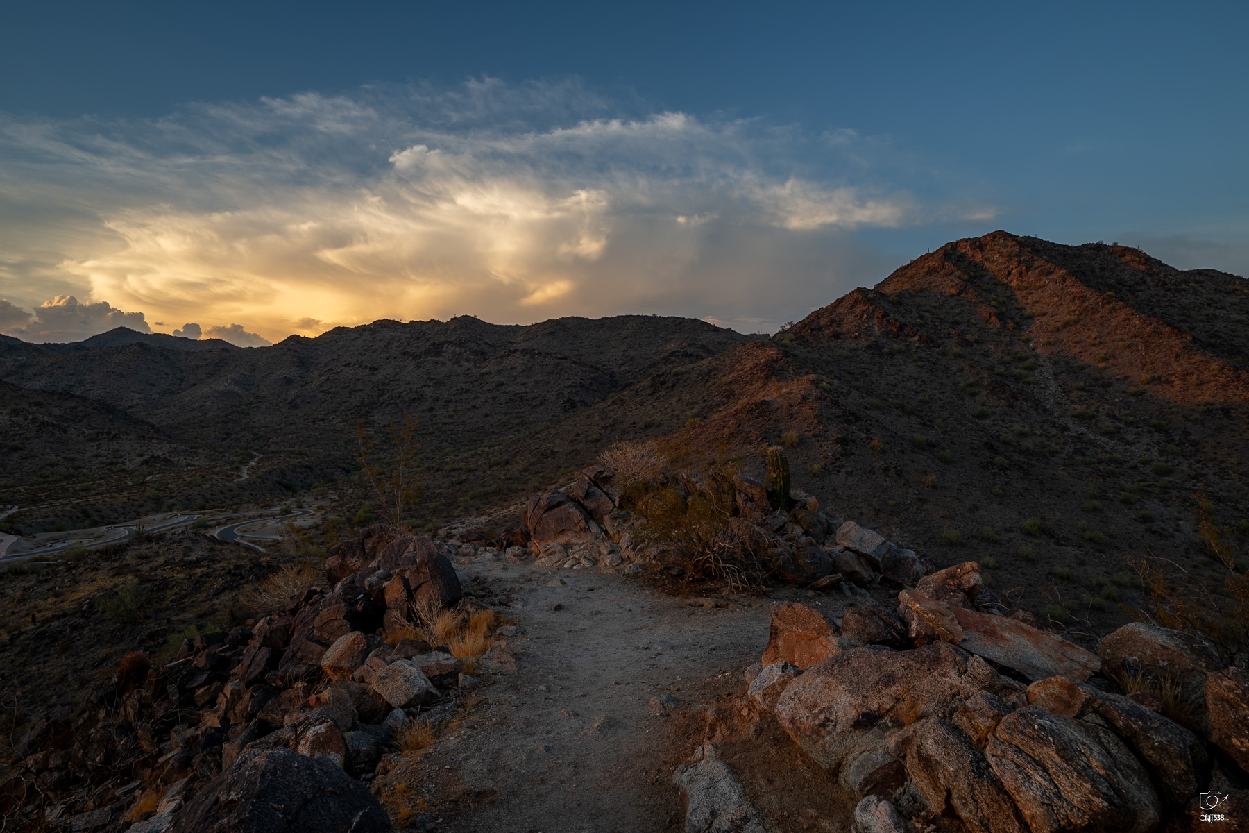 Auto-generated description: A rocky desert trail winds through rugged hills under a dramatic sky at sunset.