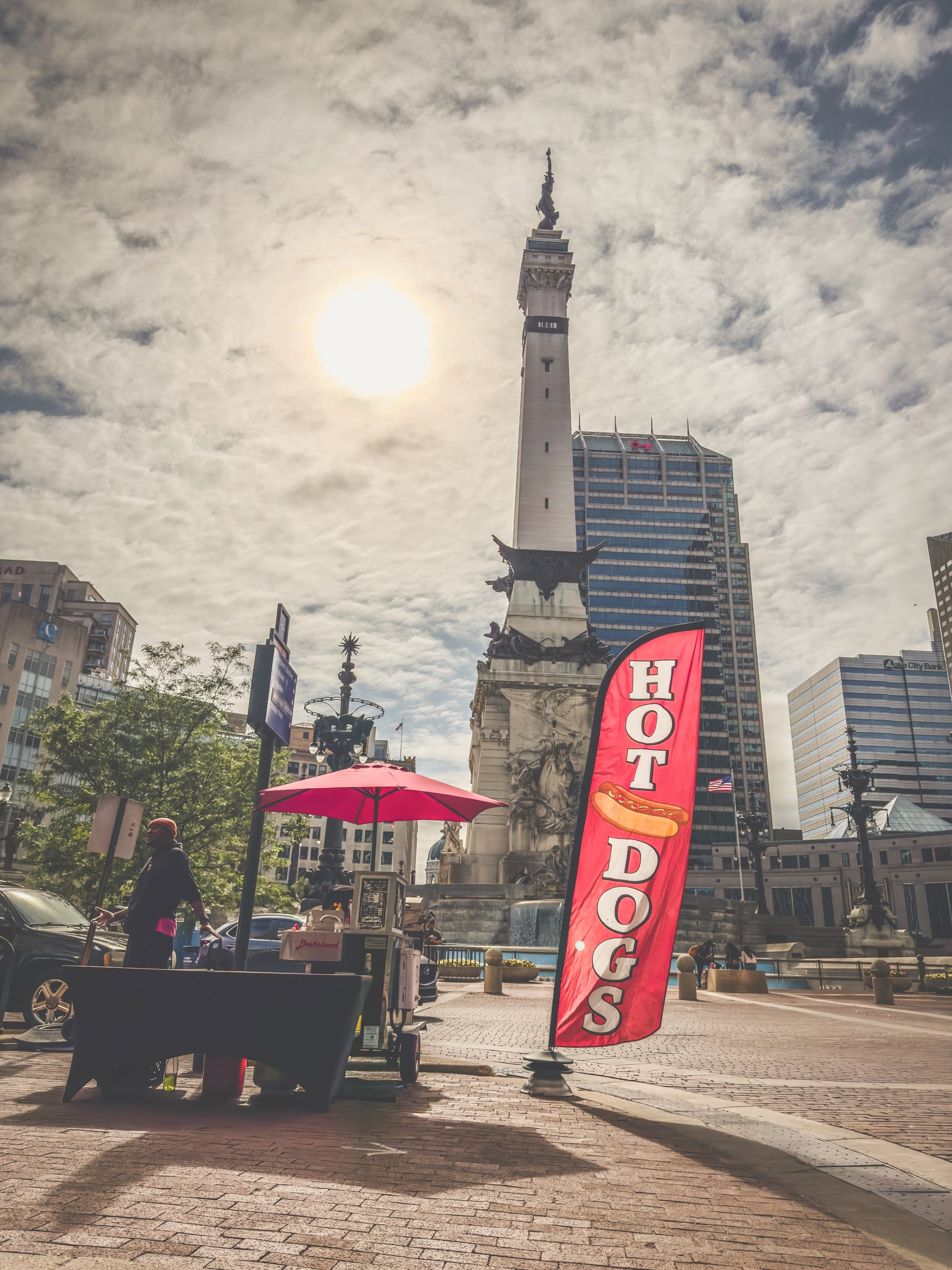 Auto-generated description: A hot dog stand with a red banner is set up in a plaza near a tall monument, surrounded by modern buildings under a cloudy sky.