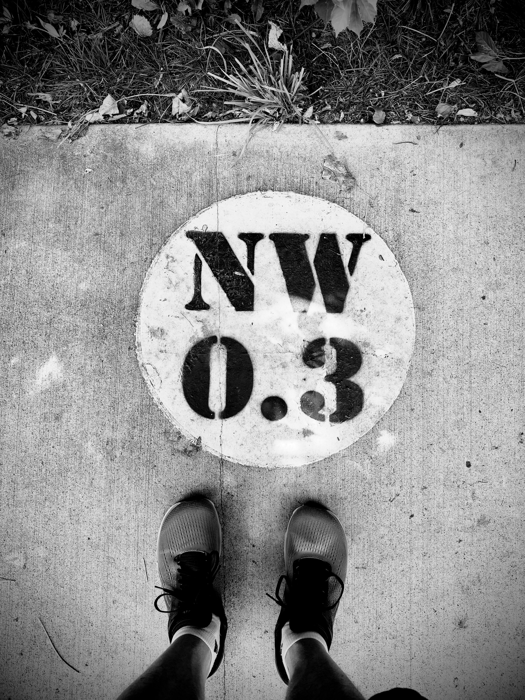 Auto-generated description: A pair of feet wearing sneakers stands on a sidewalk next to a circular marking with NW 0.3 painted on it.