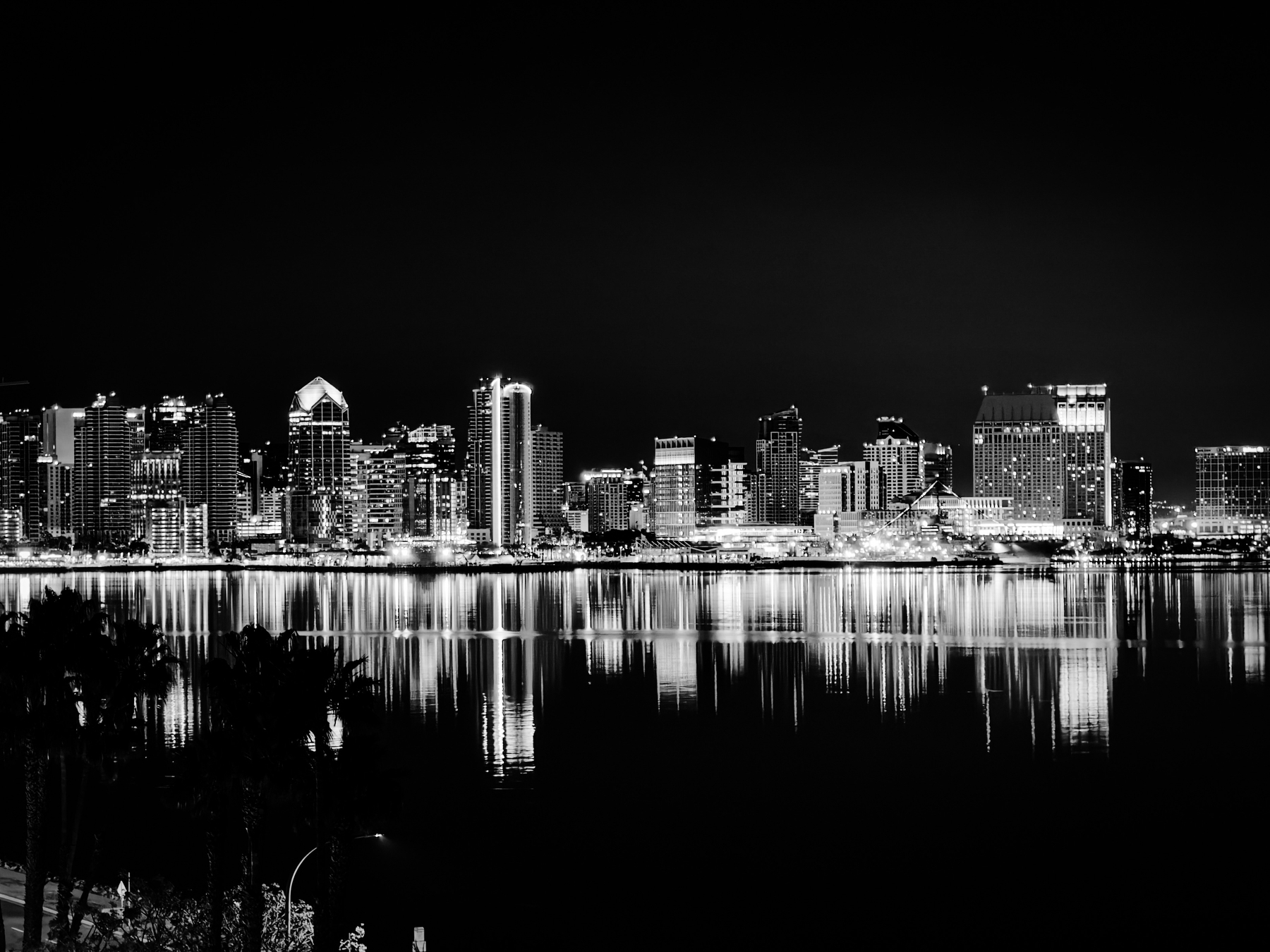 Auto-generated description: A black-and-white cityscape depicts a skyline with illuminated buildings reflecting on a calm body of water at night.