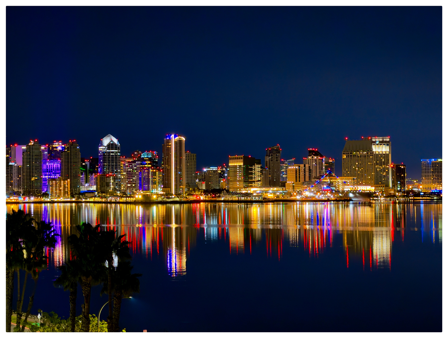 A vibrant city skyline is illuminated at night, with colorful reflections shimmering on the water.