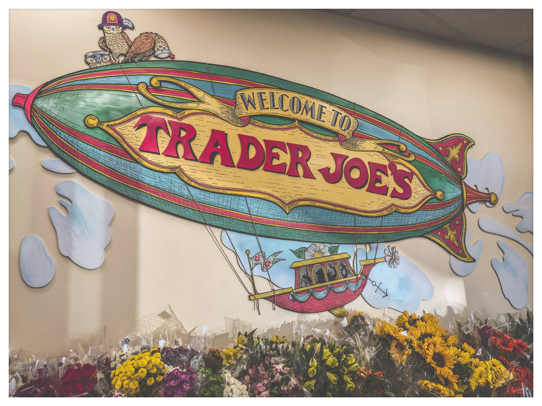 Auto-generated description: A colorful, whimsical mural featuring a blimp with the words Welcome to Trader Joe's is displayed above an array of vibrant flowers.