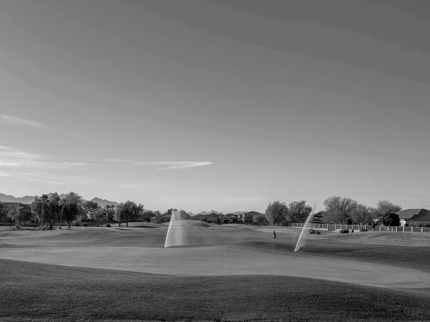 Auto-generated description: A golf course with sprinklers watering the green under a clear sky.
