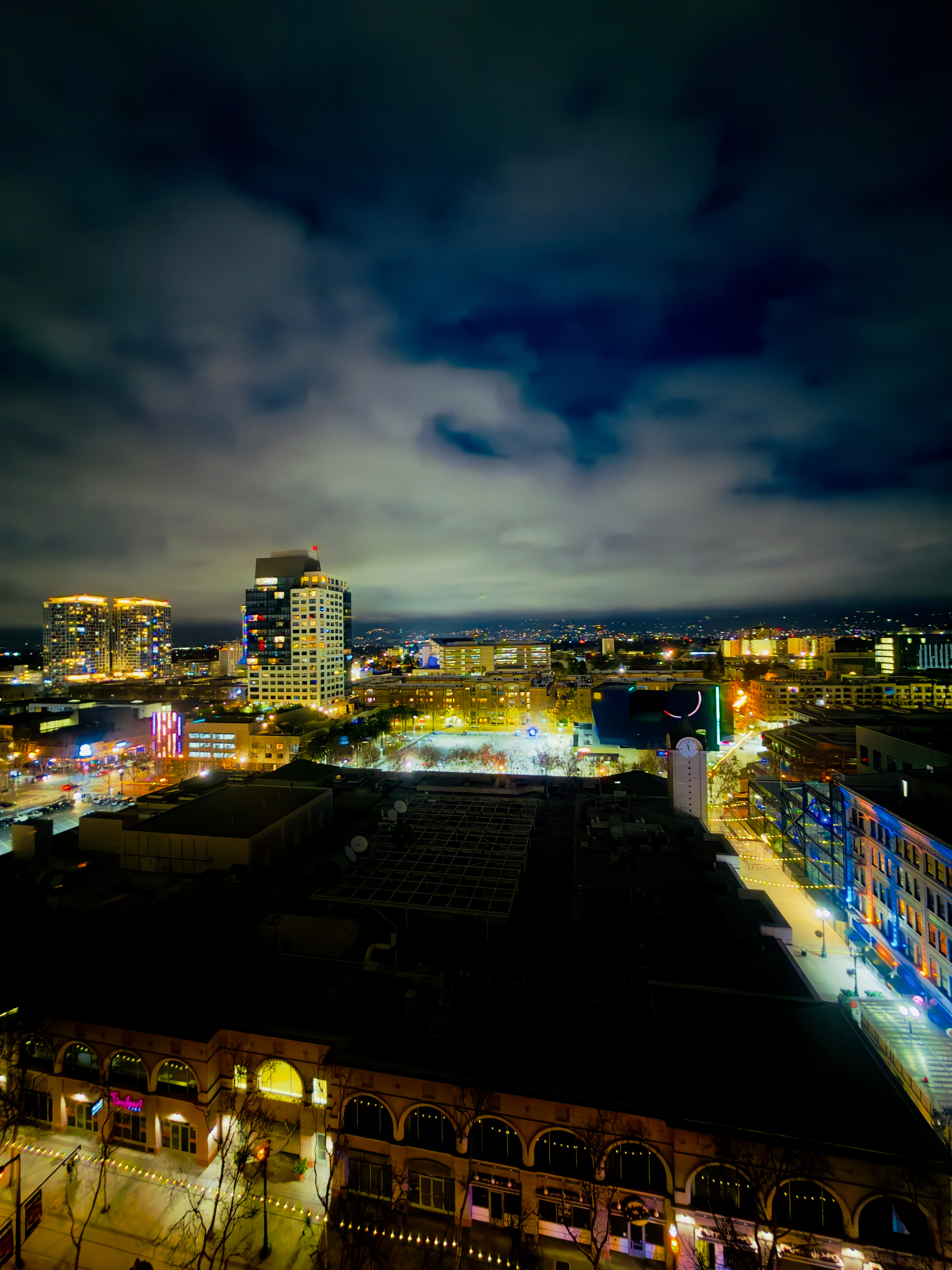 Auto-generated description: A vibrant cityscape at night showcases illuminated buildings under a partly cloudy sky.
